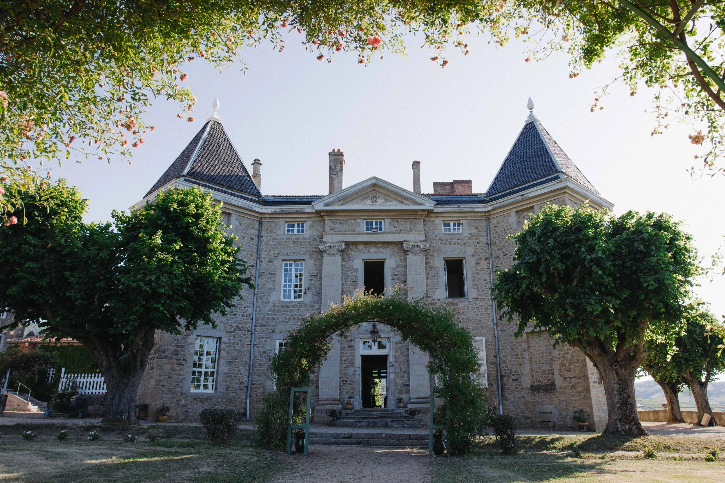 Château en pierre avec deux tours, entouré d'arbres verts, avec un chemin menant à une porte sous un arc végétal, sous un ciel clair. Mariage au chateau de lachal dans le rhone alpes à côté de Lyon. Cérémonie civil et laïque. Robe de mariées.