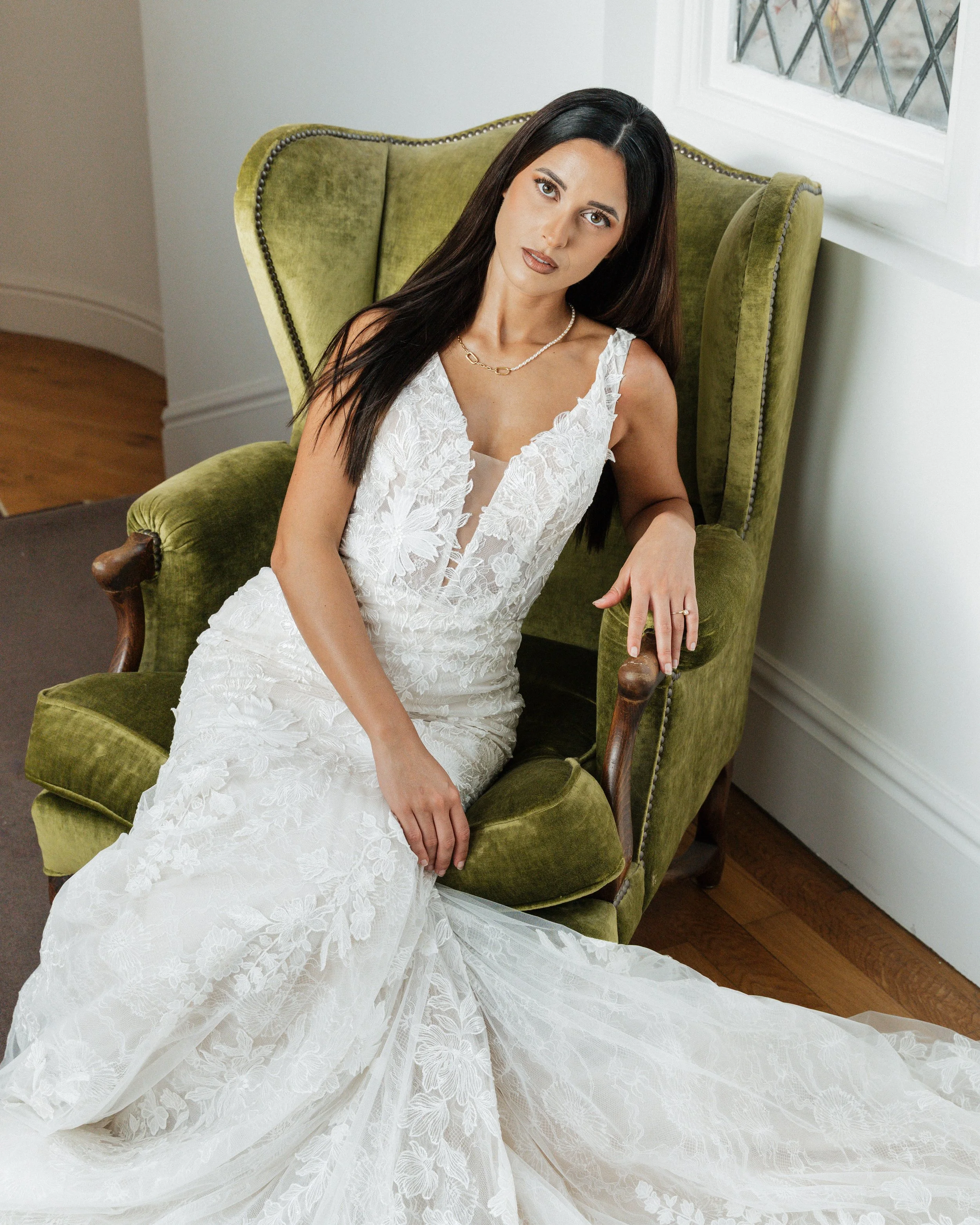 Une femme en robe de mariée blanche assise dans un fauteuil vert ancien près d'une fenêtre.  session femme mariées et robes haut de gamme, brindos, hôtel et lac,  photographe mariage bordeaux et Lyon et destination wedding