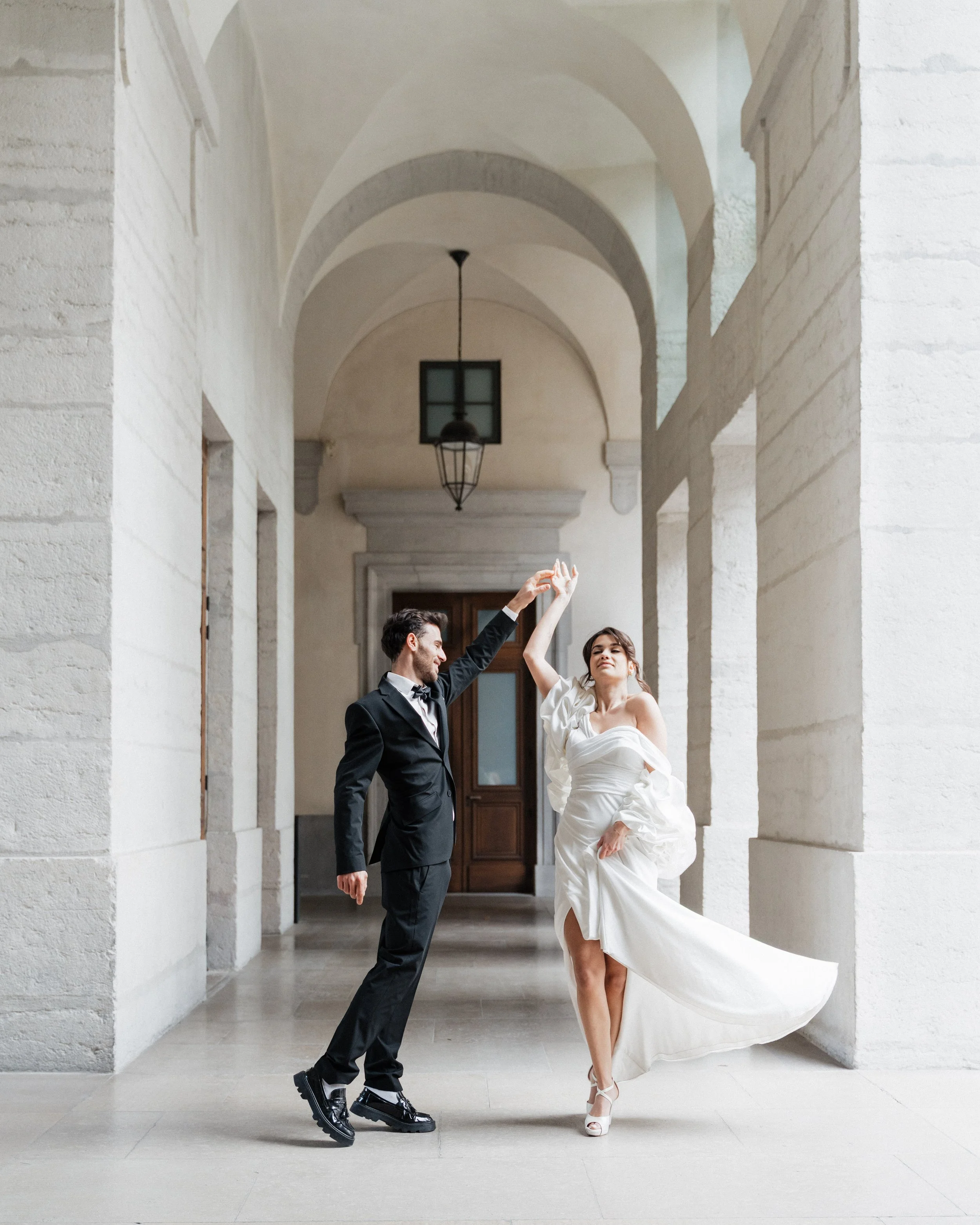 Un couple en vêtements de mariage danse dans un couloir architecturé blanc avec des arches et une lumière suspendue à Lyon 2ème à l'hôtel dieu à l'intercontinental lors d'une session engagement par un duo de photographe de mariage et de couples. 