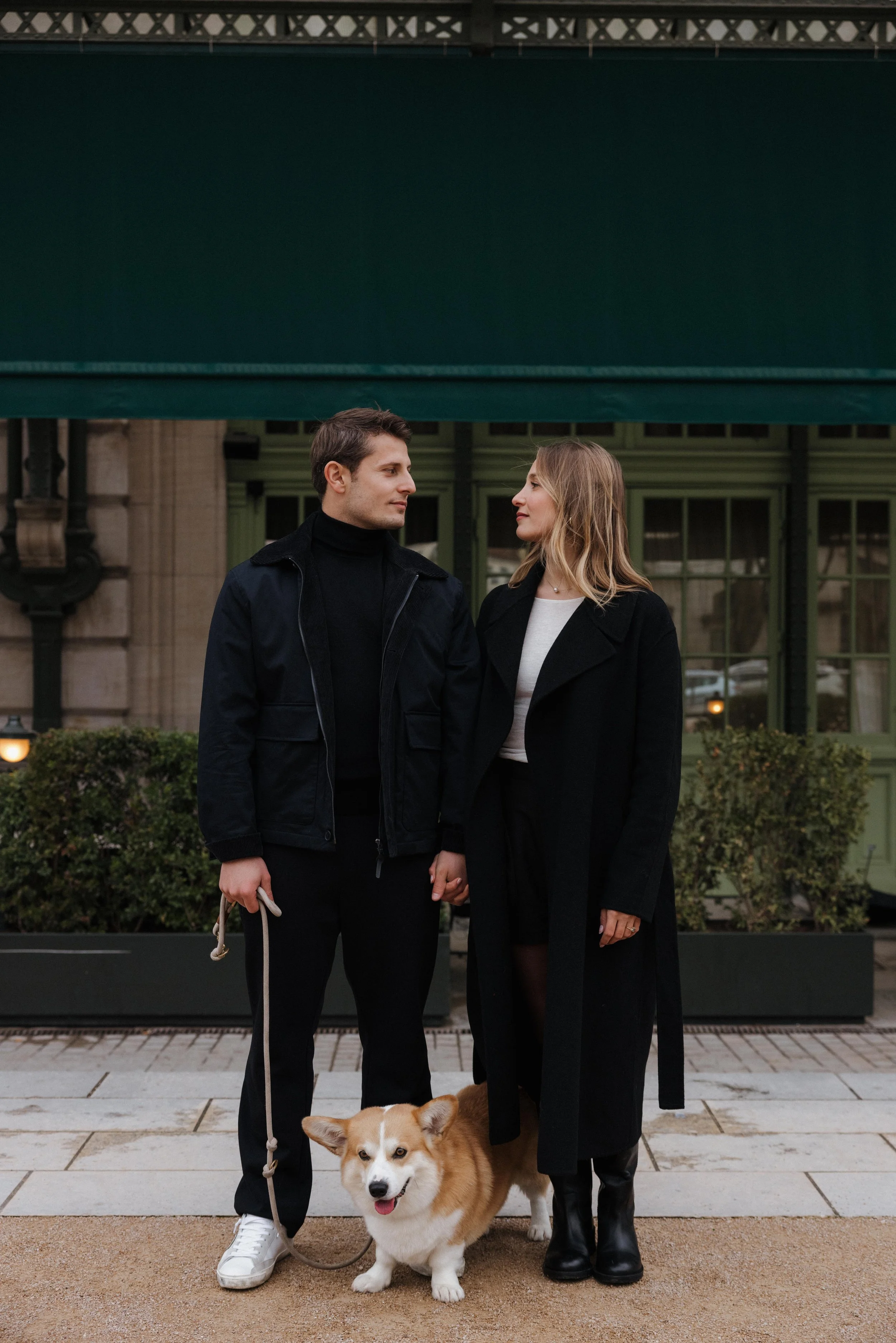 Un coupleTenant la main, accompagnée d'un chien de race Pembroke Welsh Corgi, se tient devant un bâtiment avec une façade verte et des fenêtres à carreaux, dans un contexte urbain. La femme porte un manteau noir long et des bottes, tandis que l'homme