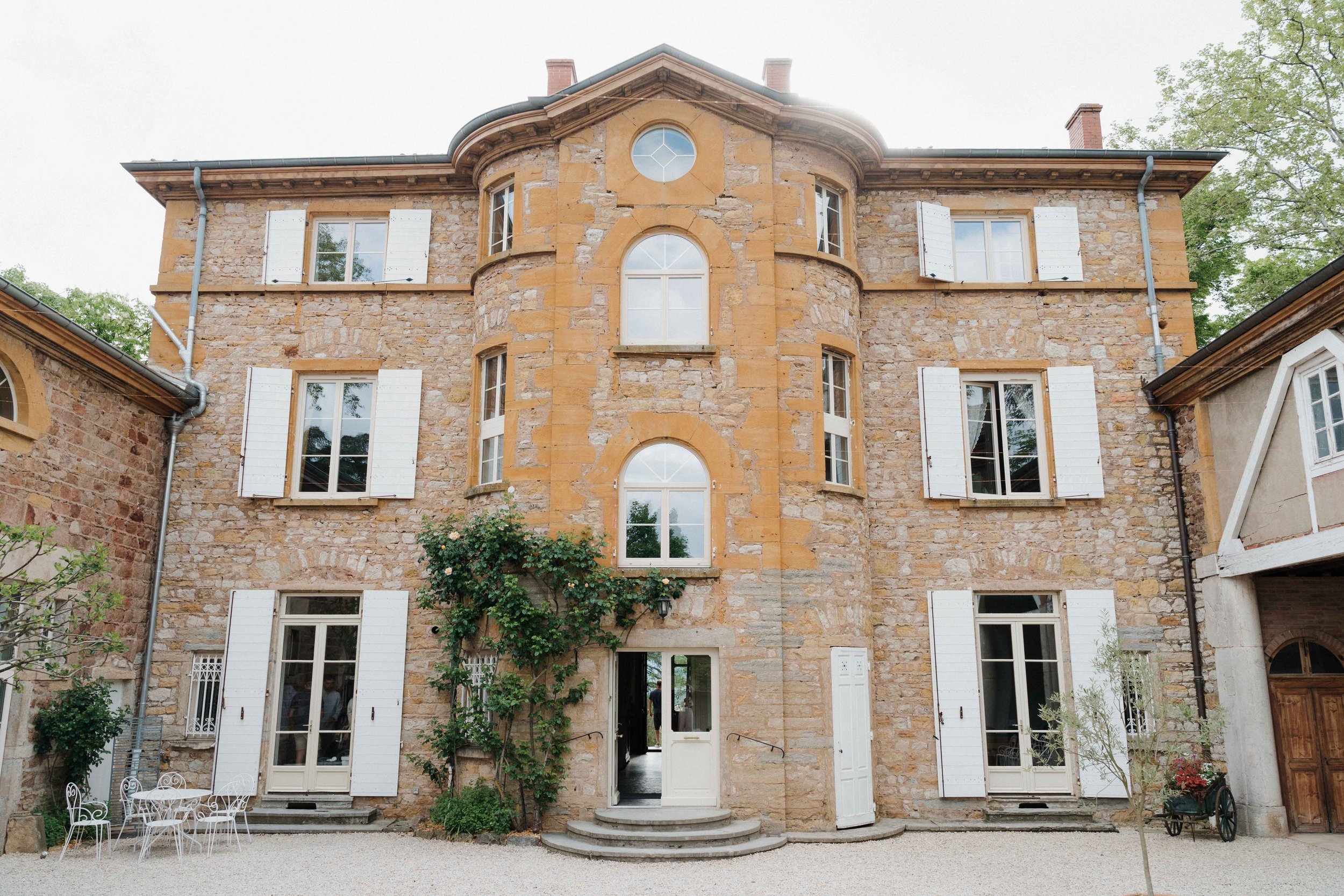 Facade en pierre d'une vieille maison avec fenêtres à volets blancs, arbres et mobilier extérieur. Domaine de tourieux à Savigny dans le beaujolais à Lyon. Mariage cérémonie laïque, photographe Lyon haut de gamme, duo photographe, argentique.