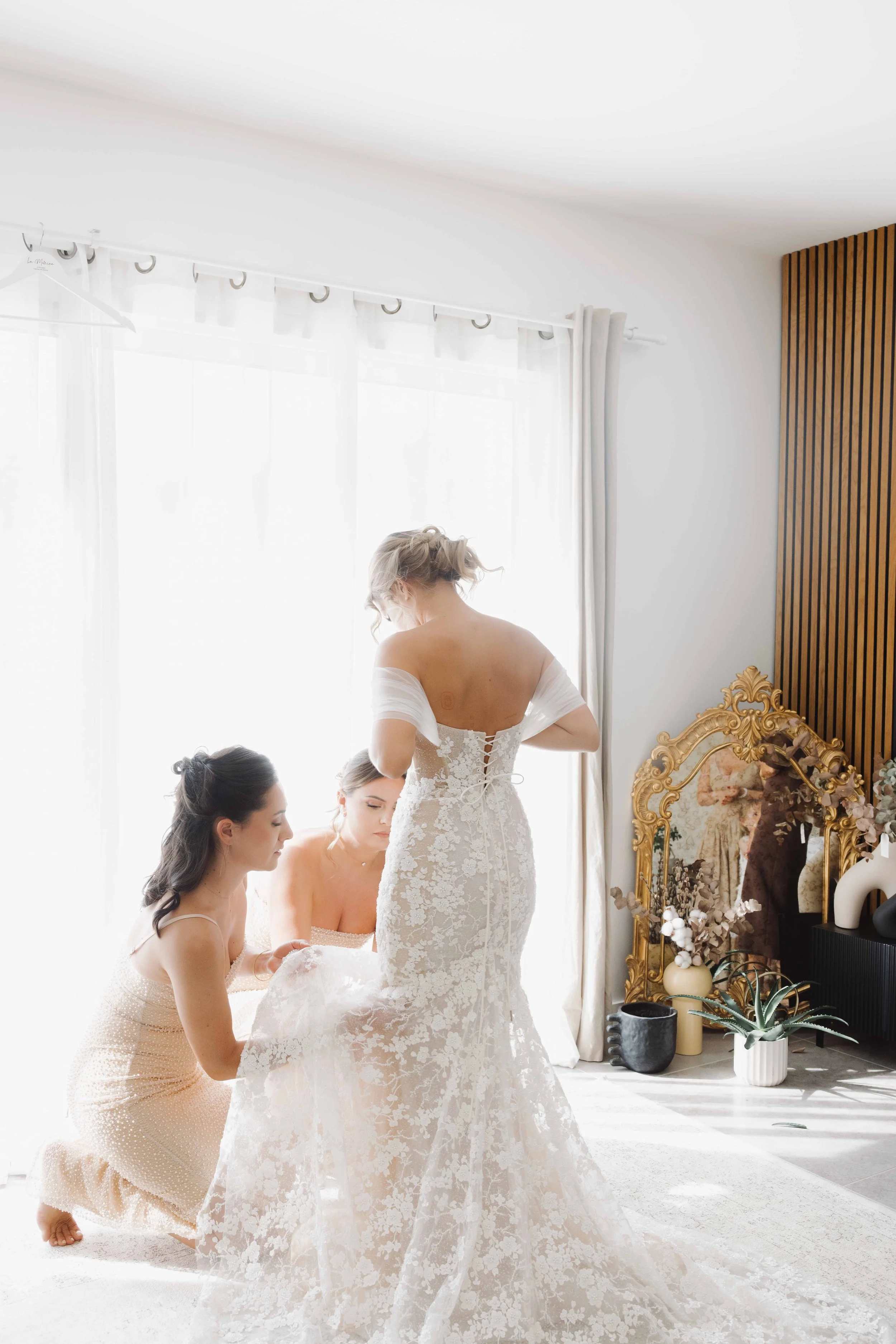Femme en robe de mariée se préparant avec deux amies dans une pièce lumineuse, décorée avec un grand miroir doré, des plantes et des vases. Photographe mariage Isère, ferme du rocher, cérémonie civil et laïque. Bordeaux et Lyon et destination wedding