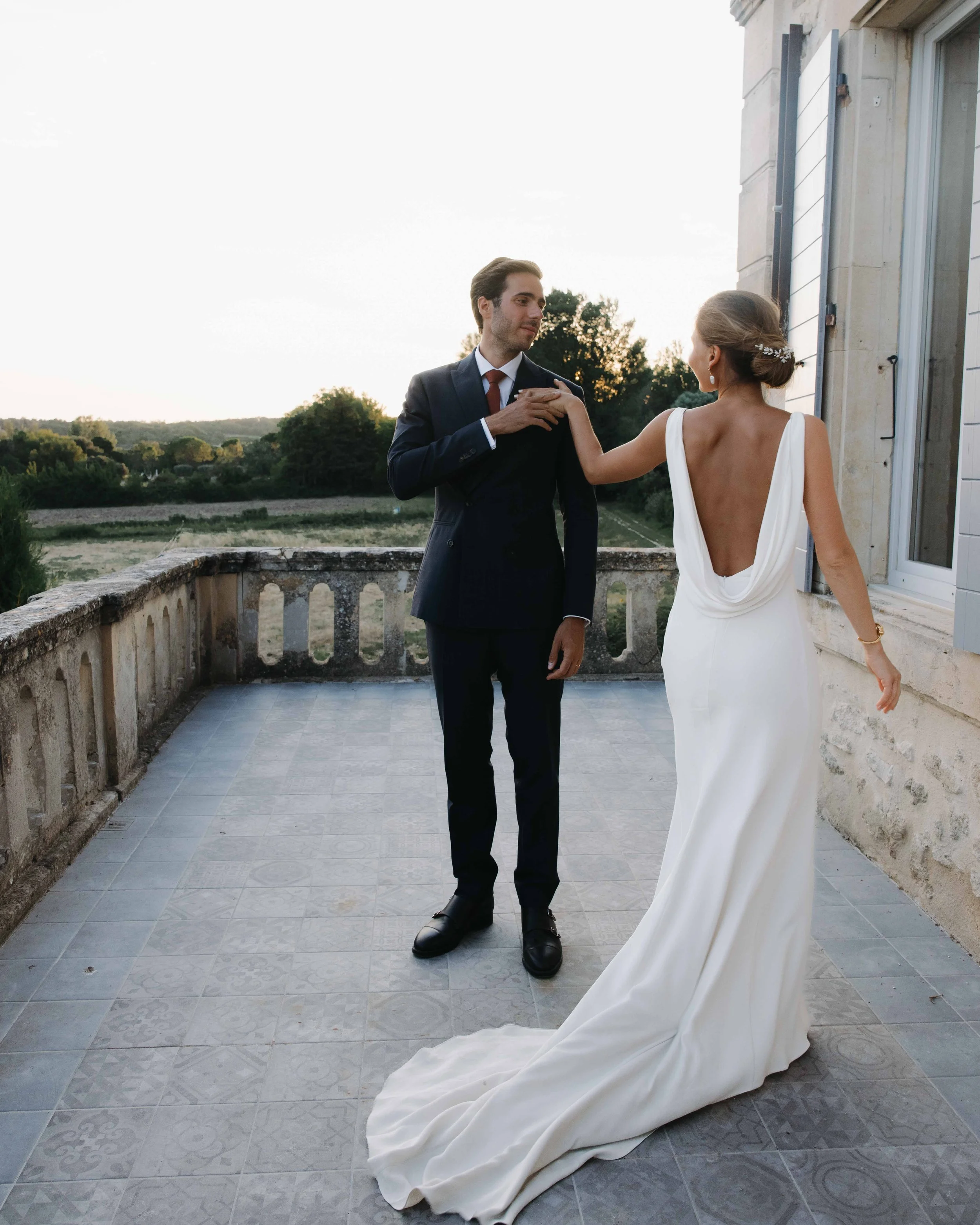 Un couple en costume de mariage sur une terrasse extérieure, la femme en robe blanche dos nu, le homme en costume foncé, échangeant un moment sentimental au coucher du soleil. Domaine du bijoutier à grignan, drome provençal, session couple, danse 