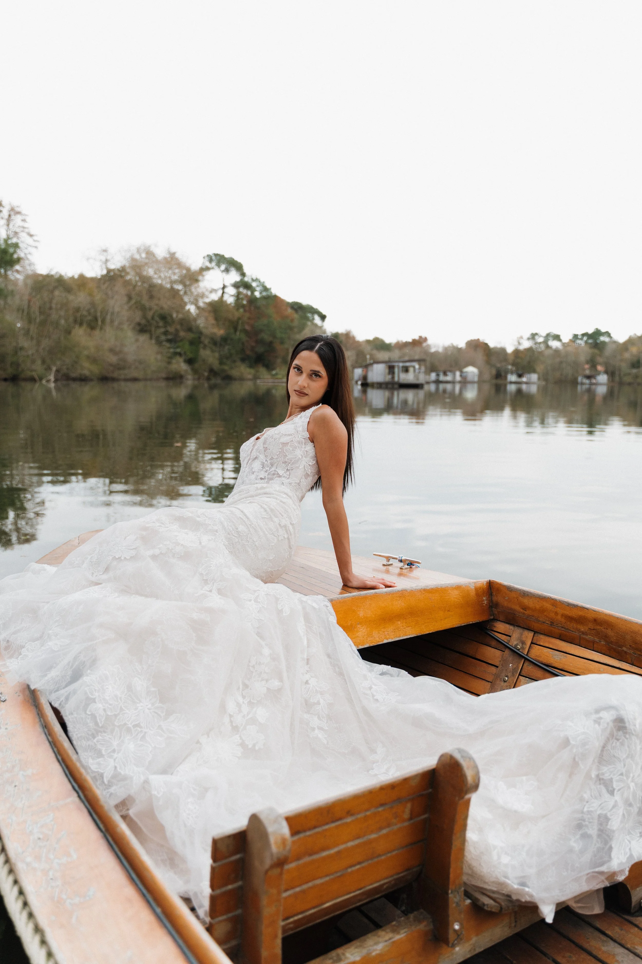 Une femme en robe de mariée blanche assise sur une embarcation en bois sur un lac calme, avec des arbres en arrière-plan  session femme mariées et robes haut de gamme, brindos, hôtel et lac,  photographe mariage bordeaux et Lyon et destination weddin
