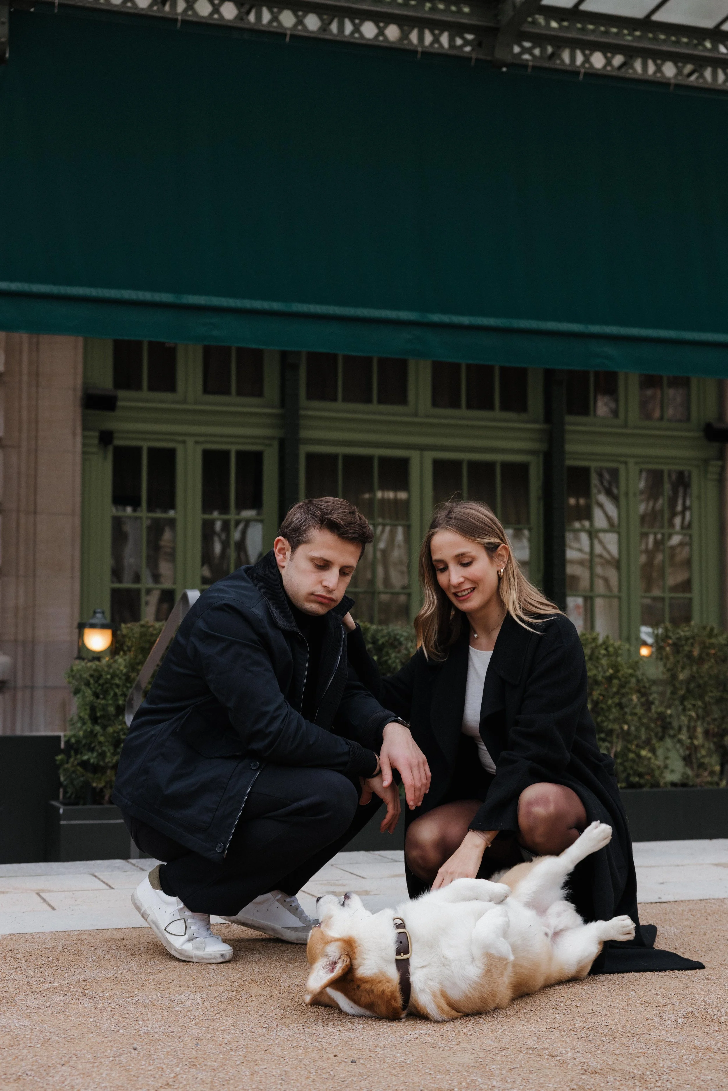Un couple joue avec un chien dans un espace urbain, sous une grande structure métallique. La femme sourit, le chien roux et blanc est allongé sur le sol. session engagement couple Lyon 6eme, brotteaux, photographe mariage bordeaux et Lyon