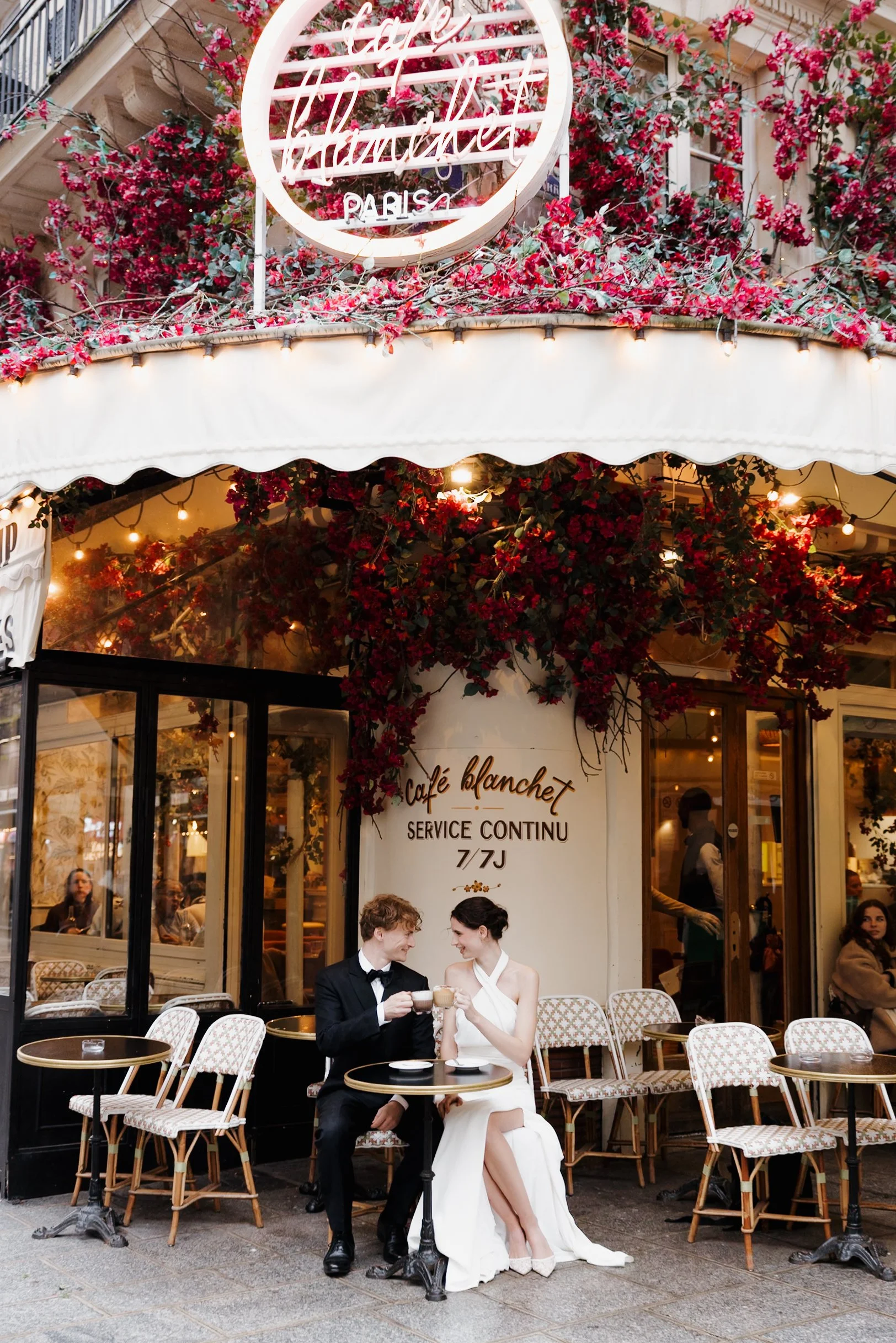 Un couple habillé élégamment, un homme en costume noir et une femme en robe blanche, assis à une table de café à l'extérieur devant un café parisien, en train de boire ensemble. Session engagement café parisien, photographe mariage paris 