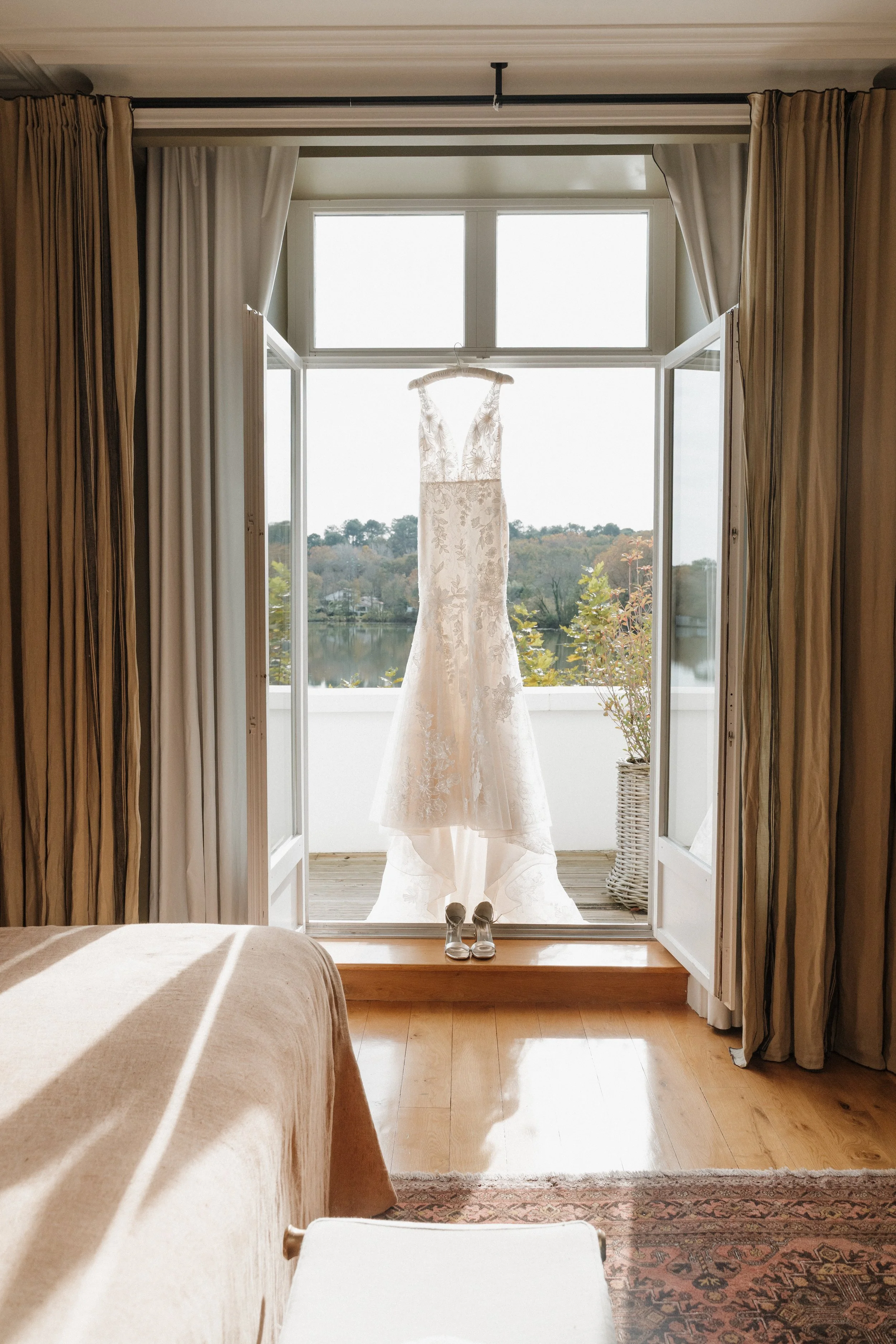 Robe de mariage blanche suspendue à un crochet, vue depuis l'intérieur d'une chambre, sur un balcon avec vue sur un lac et des arbres, avec une paire de chaussures à talons en premier plan.  session femme mariées et robes haut de gamme, brindos