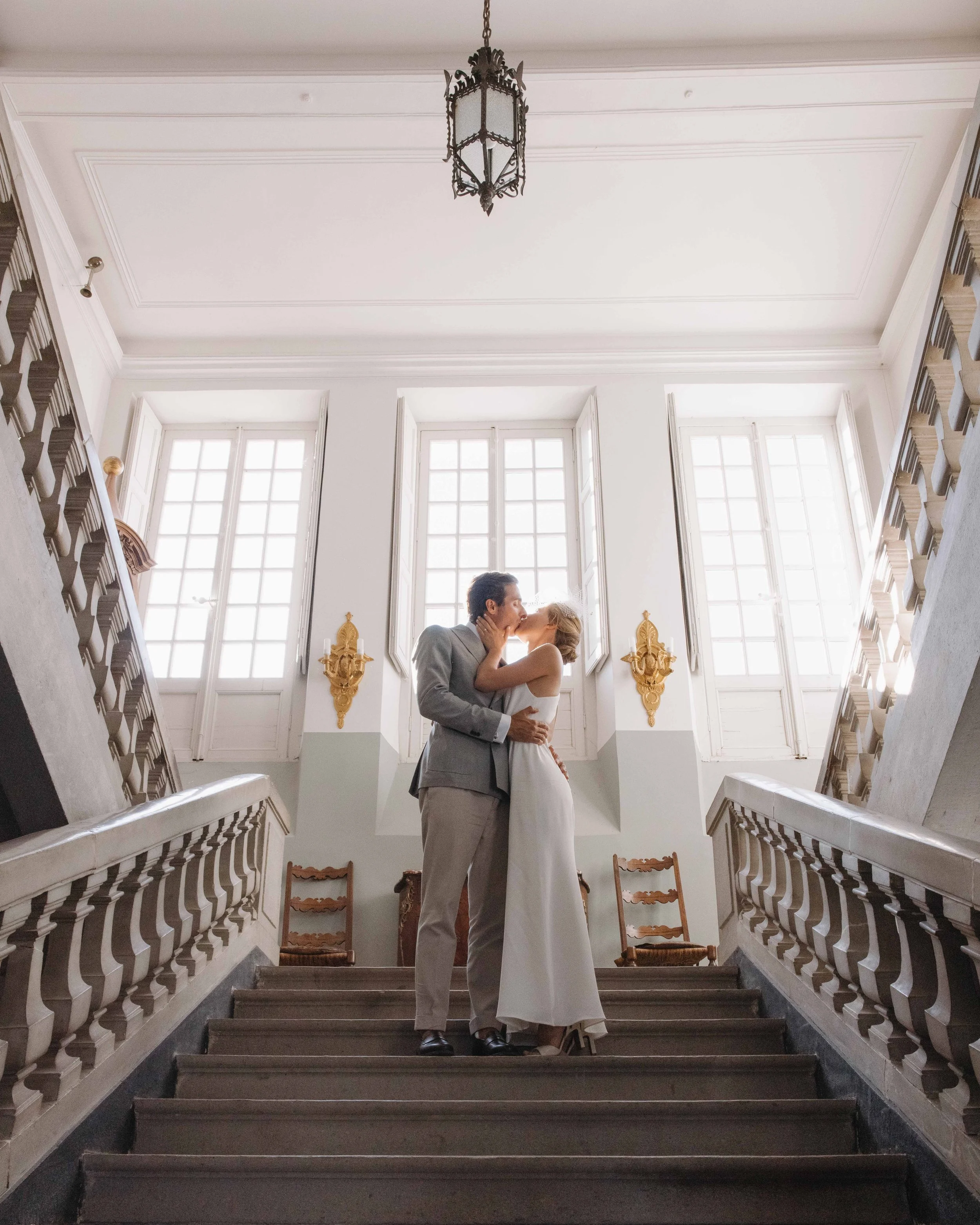 Un couple de mariés s'embrassant au sommet d'un escalier dans une salle lumineuse avec de grandes fenêtres. Mariage au chateau de lachal dans le rhone alpes à côté de Lyon. Cérémonie civil et laïque. Robe de mariées et costume homme. Photographe duo 