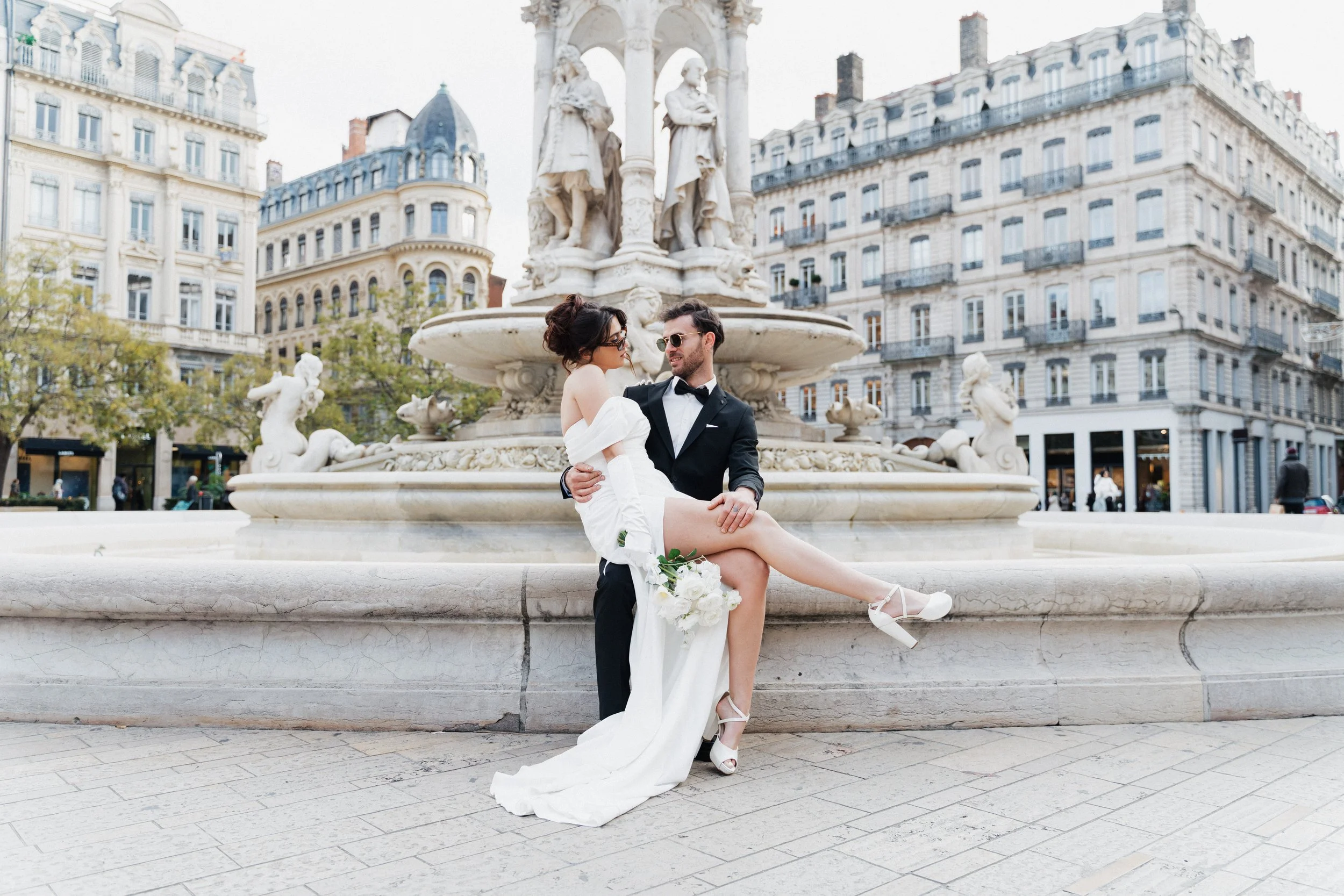 Un couple de mariés posant devant une fontaine historique à Paris, avec des bâtiments haussmanniens en arrière-plan, l'homme en costume noir et la femme en robe blanche de mariée. à Lyon,place des jacobins, photographe duo haut de gamme mariage Lyon 