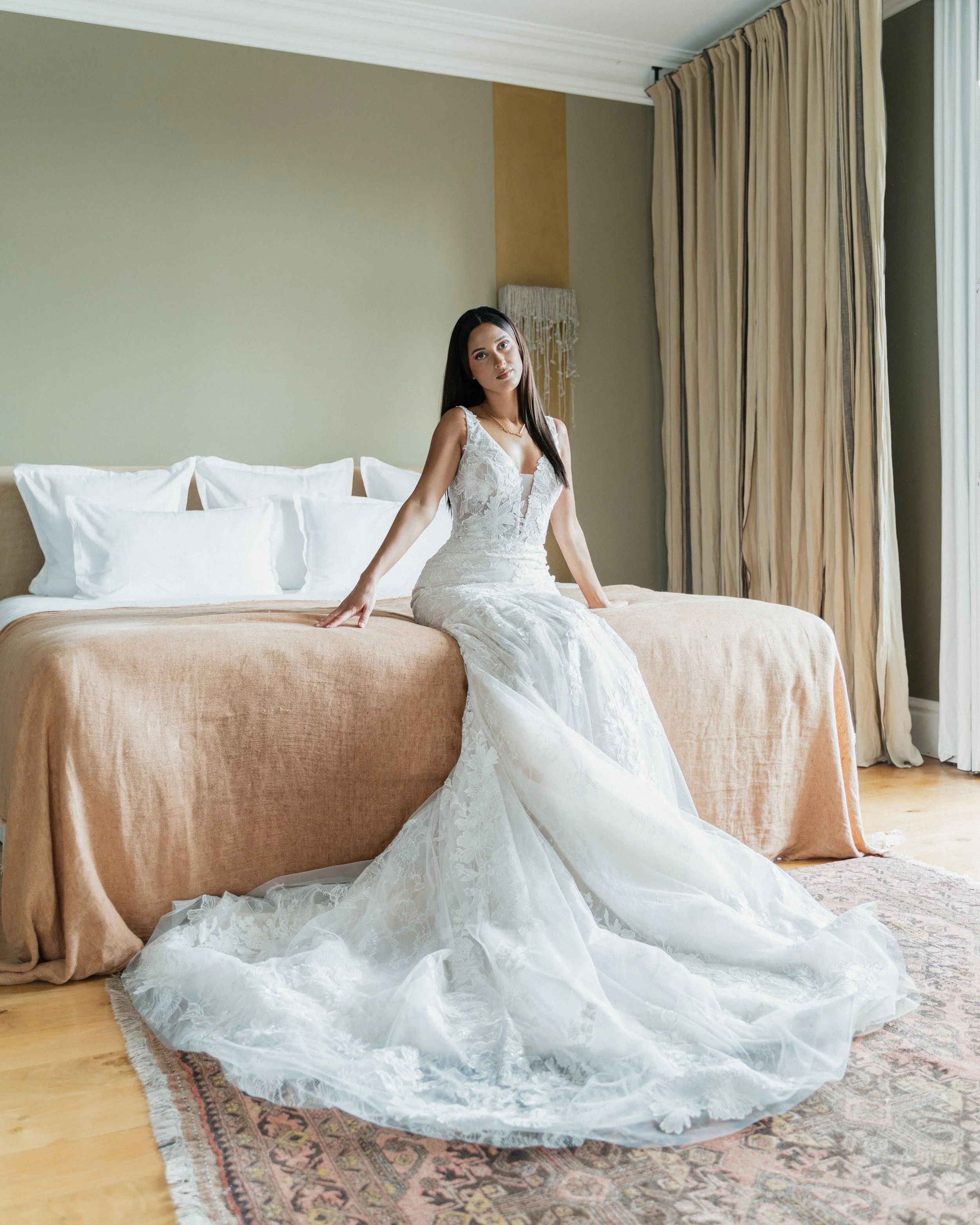Une femme en robe de mariée assise sur un lit dans une chambre lumineuse avec rideaux beige et mur vert pâle.  session femme mariées et robes haut de gamme, brindos, hôtel et lac,  photographe mariage bordeaux et Lyon et destination wedding