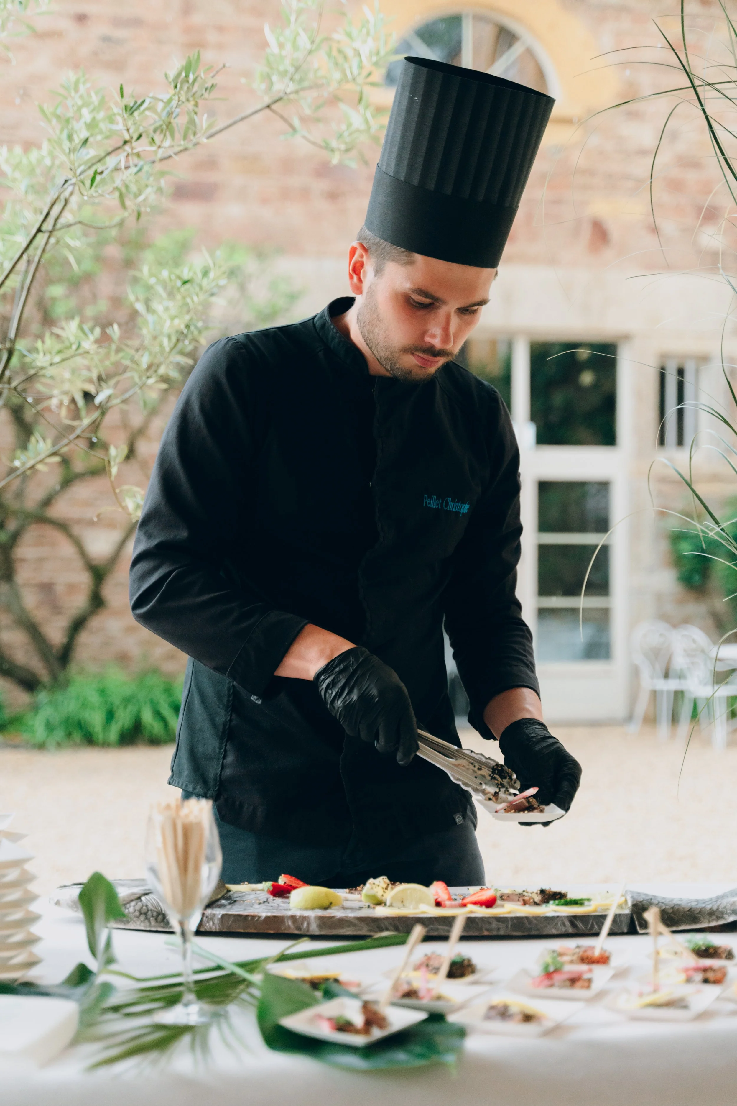 Chef en noir avec toque noire, préparant des plats lors d'un service de restauration en plein air. Domaine de tourieux à Savigny dans le beaujolais à Lyon. Mariage cérémonie laïque, photographe Lyon haut de gamme, duo photographe, argentique.
