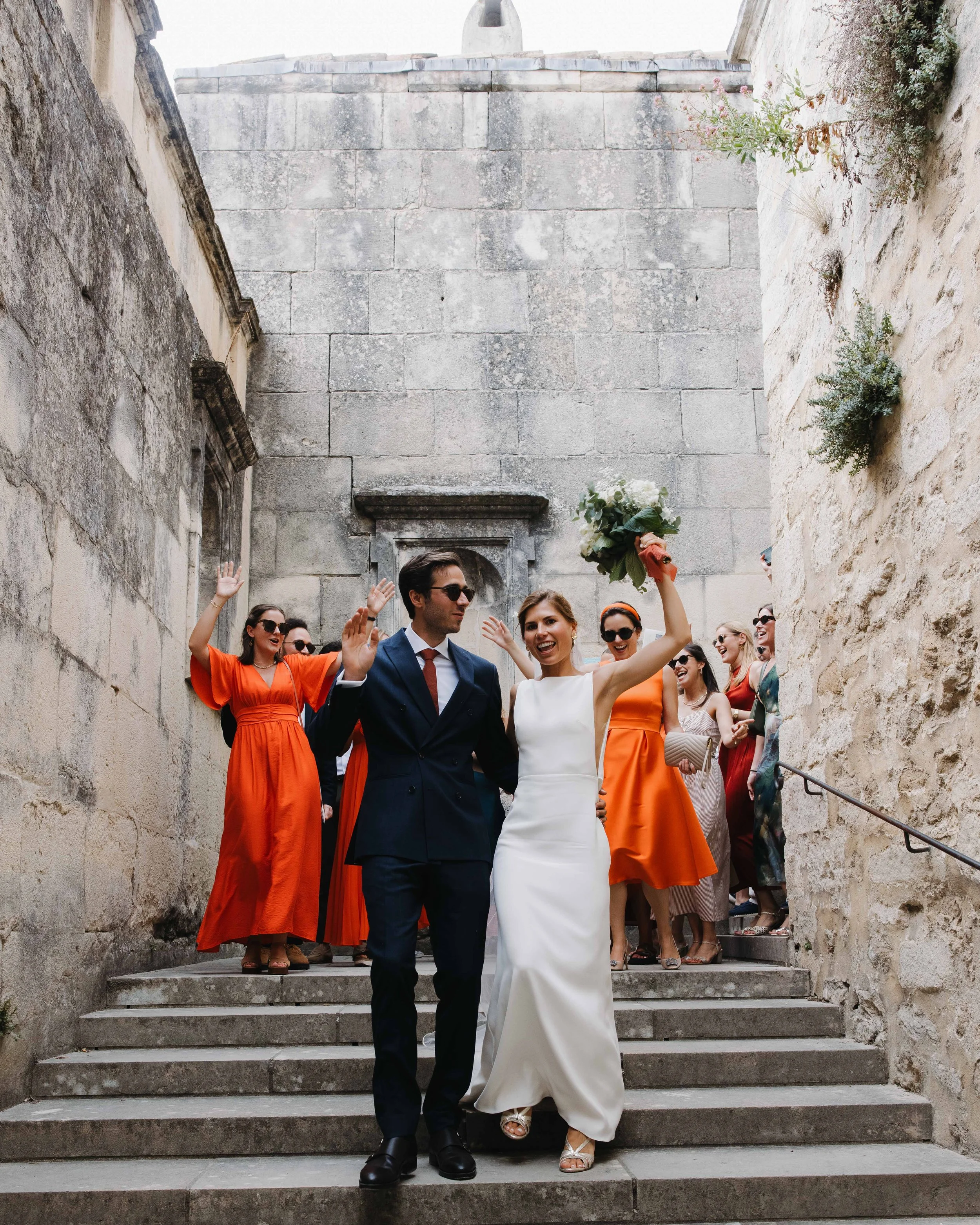 Un couple de mariés descendant des escaliers avec des amis, en célébration, la femme tenant un bouquet de fleurs, tous souriants et en tenue de mariage dans une vieille cité. Domaine du bijoutier à grignan, drome provençal, coucher de soleil 