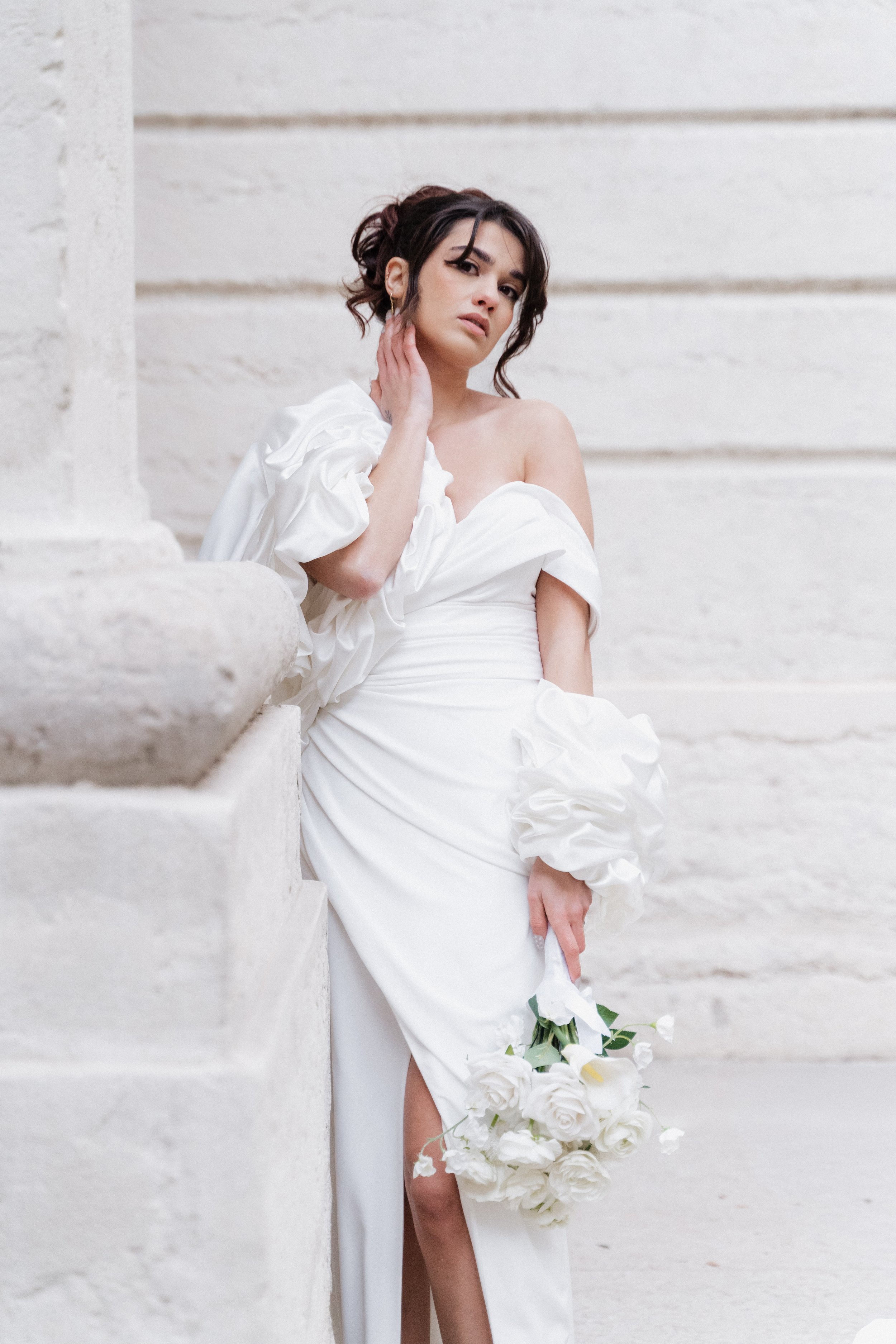 Une femme en robe de mariée blanche tient un bouquet de roses blanches et pose contre un fond de mur en pierre beige à l'hôtel dieu à Lyon 2ème, à l'intercontinental, session engament.