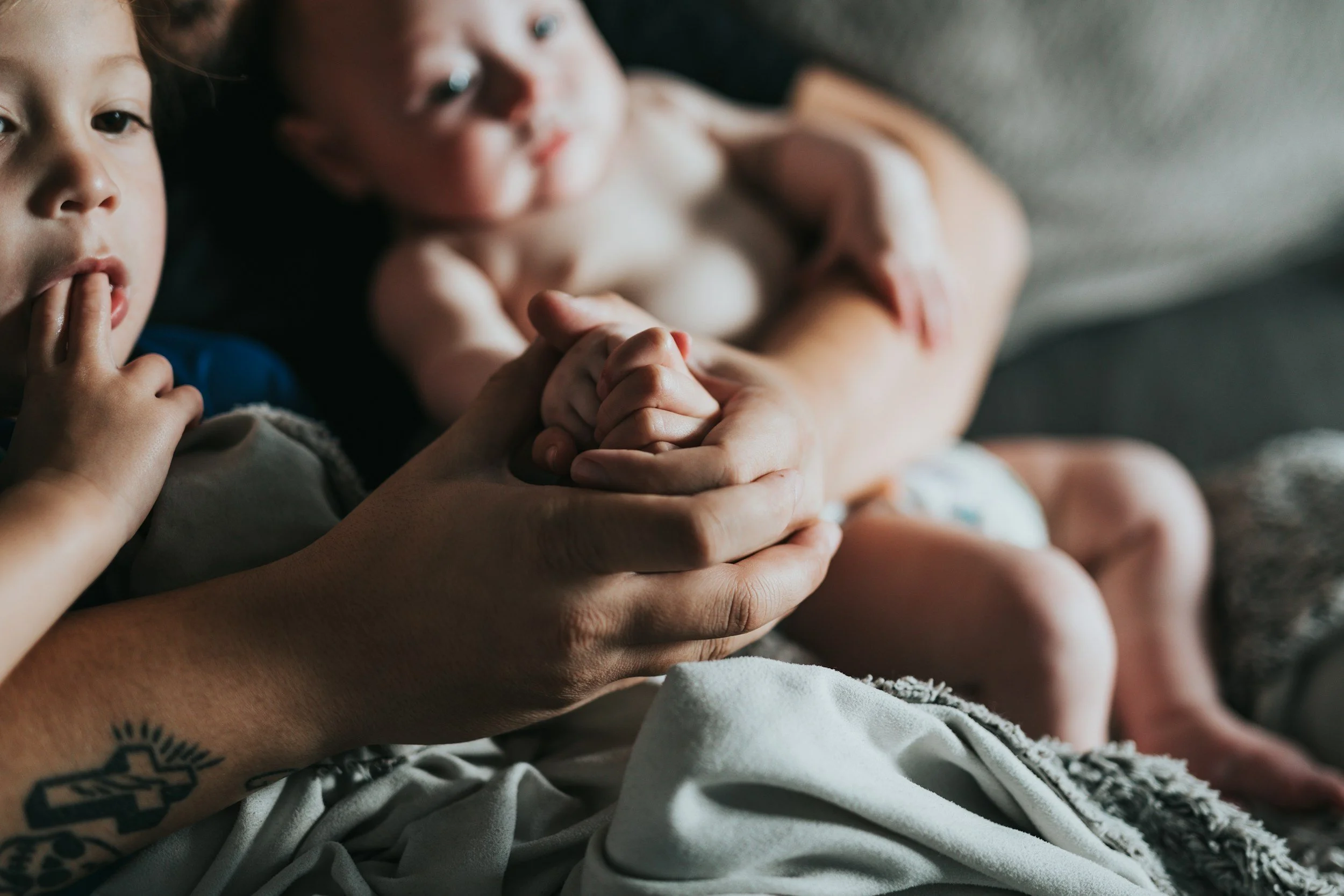 A close-up of a parent's hand holding two babies' hands, with one sucker's hand visible. The scene appears intimate and tender, set on a soft surface with a gray background.