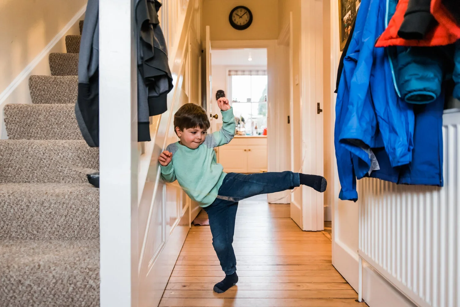 A young boy is playfully kicking his leg in a hallway, appearing to balance or pose with one arm raised. The hallway has a staircase on the left side and hanging jackets on the right side. A wall clock is mounted on the wall above, and a bright room with a window is visible at the end of the hallway.