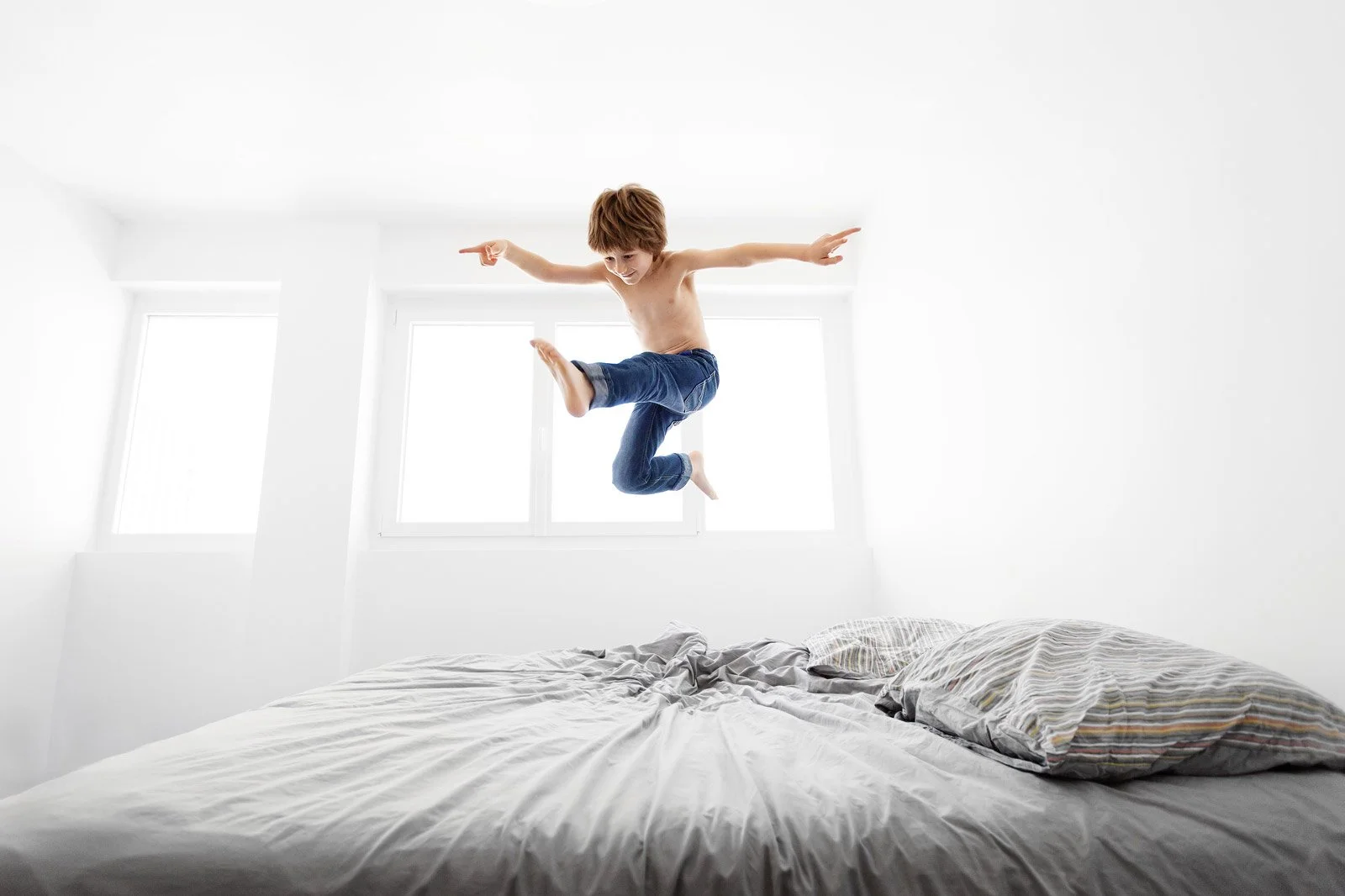 A young boy with brown hair jumping on a bed in a white room with large windows, wearing only jeans.