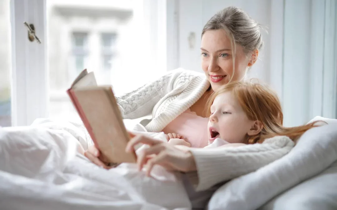 A woman and a young girl sitting in bed, reading a book together, with sunlight coming through the window.