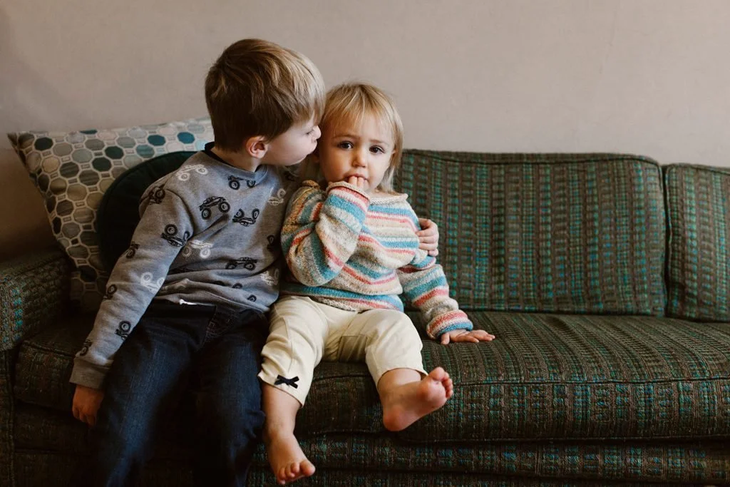 Two young children, a boy and a girl, sitting on a patterned sofa in a home setting. The boy is leaning in to kiss the girl on the cheek while the girl looks at the camera with her finger in her mouth.