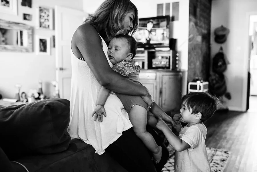 A woman holding a sleeping baby on her lap while a young boy stands next to her, touching her leg in a cozy living room.