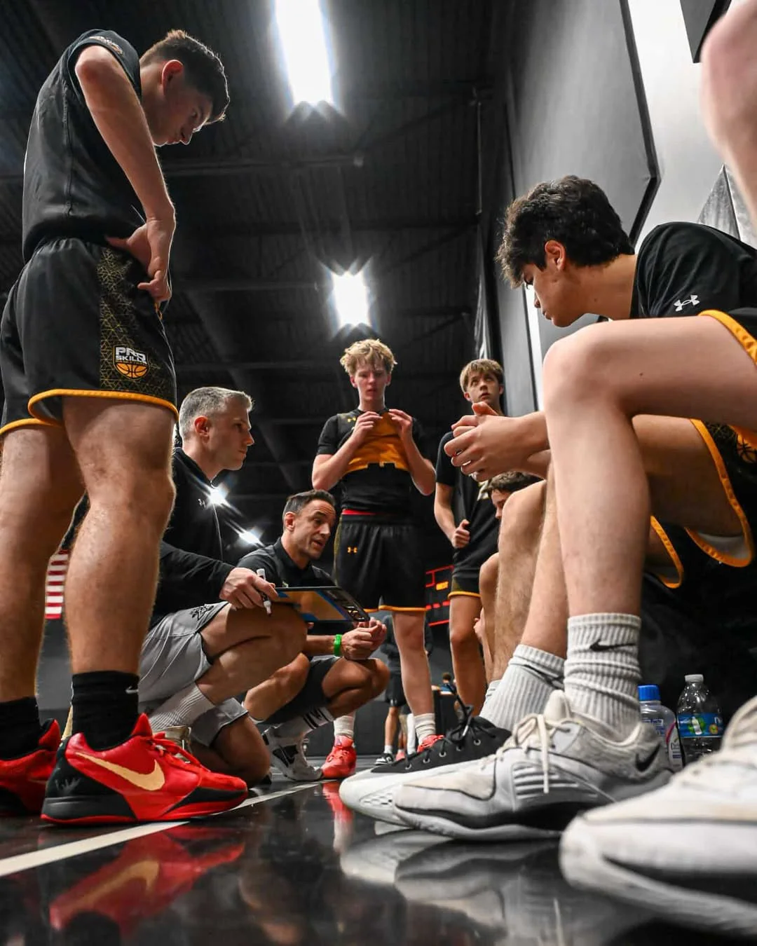 Basketball team coach giving instructions to players during a timeout in an indoor gym.