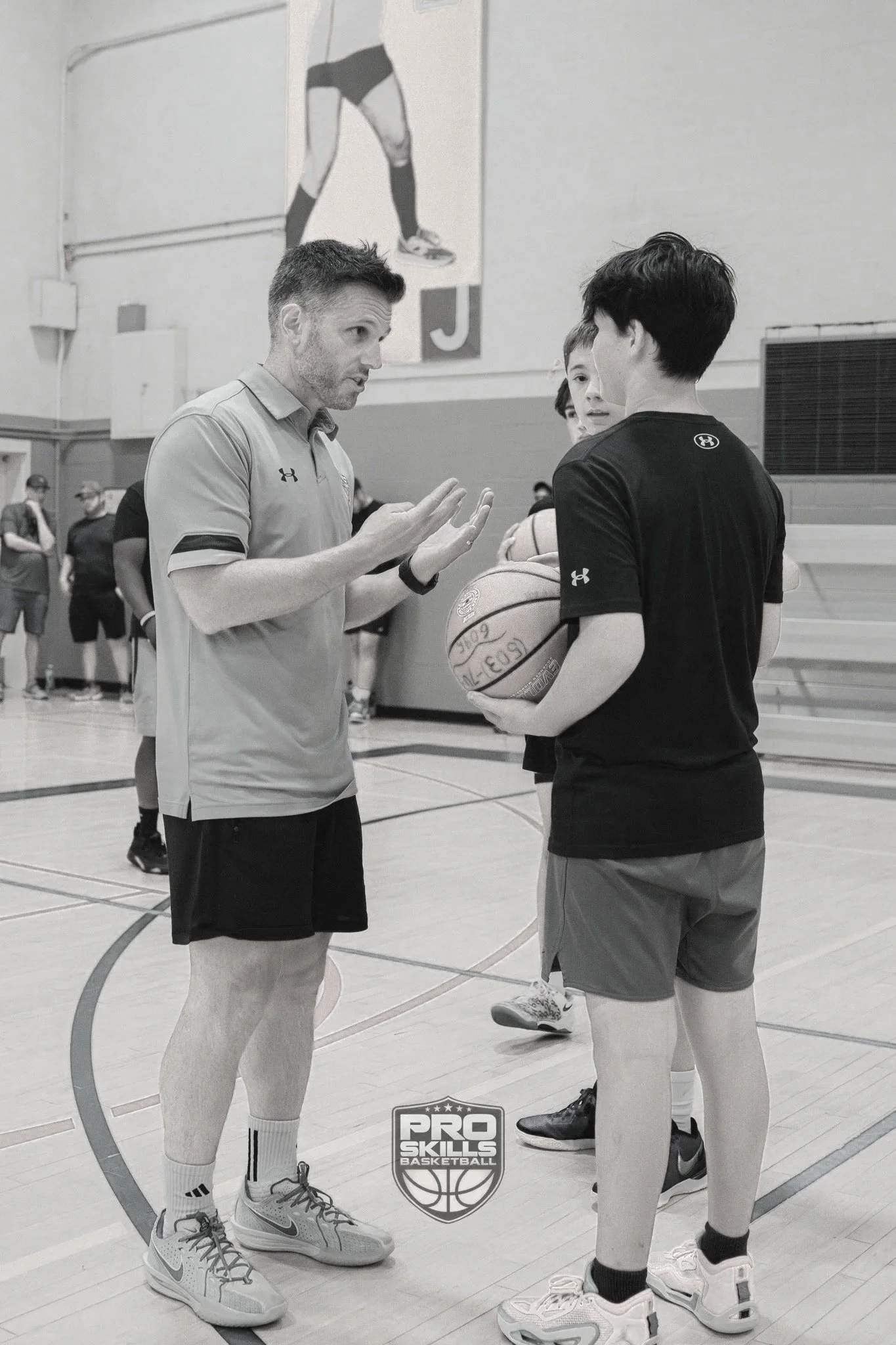A basketball coach giving instructions to a young player holding a basketball during a practice session in a gymnasium.