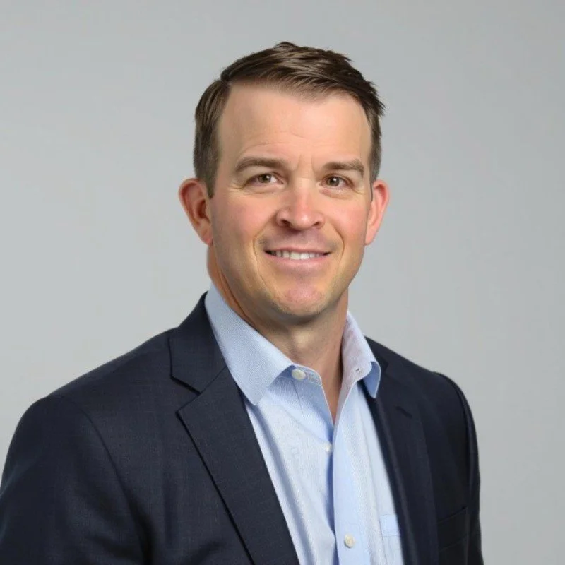 A professional headshot of a man with short brown hair, wearing a dark suit jacket and a light blue dress shirt, smiling against a plain light gray background.