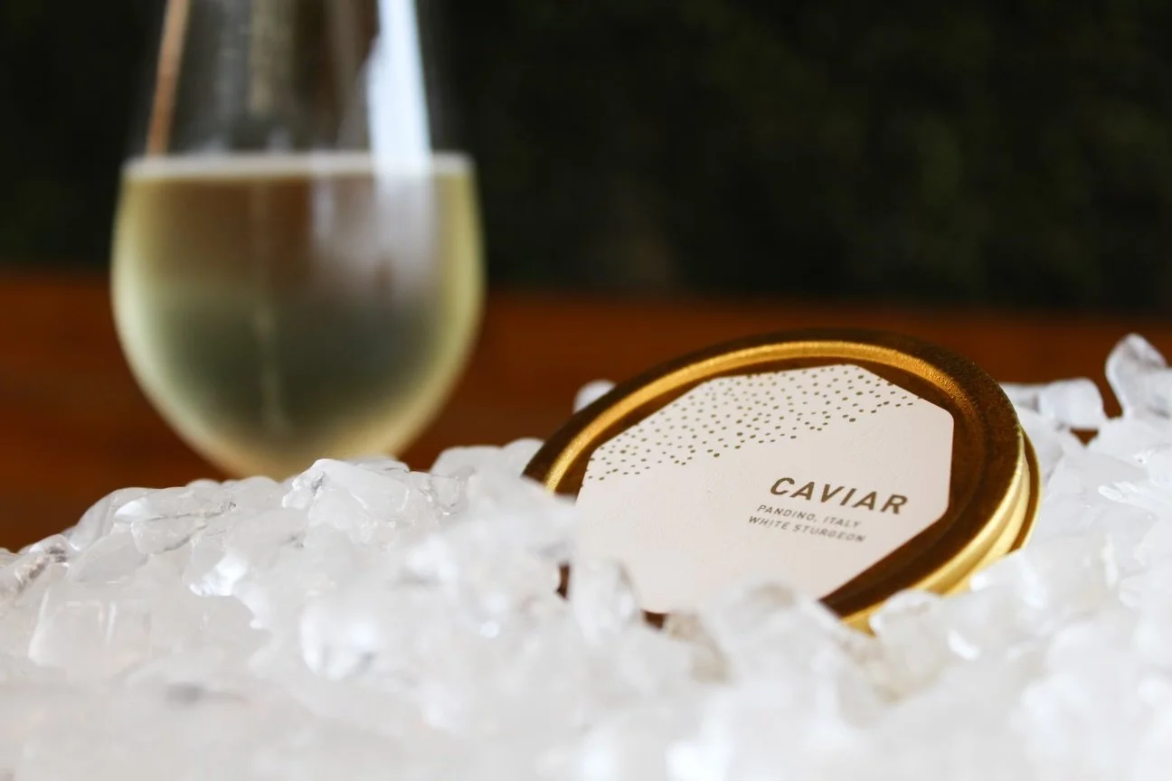 A container of Island creek caviar in a gold and white tin surrounded by ice, with a glass of white wine in the background.