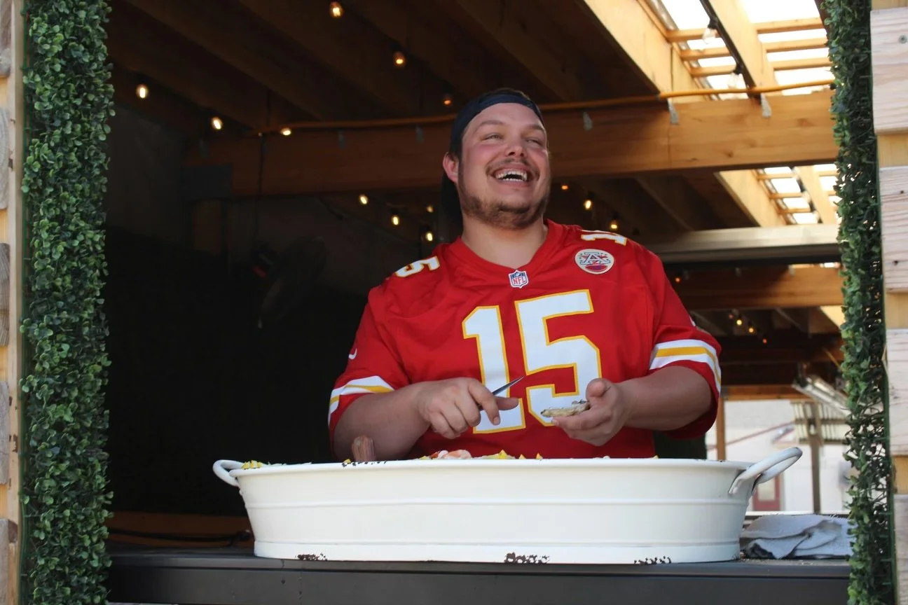 A man wearing a red Kansas City Chiefs football jersey with the number 15, smiling and holding a serving utensil, standing behind a large white serving dish inside a wooden outdoor structure.