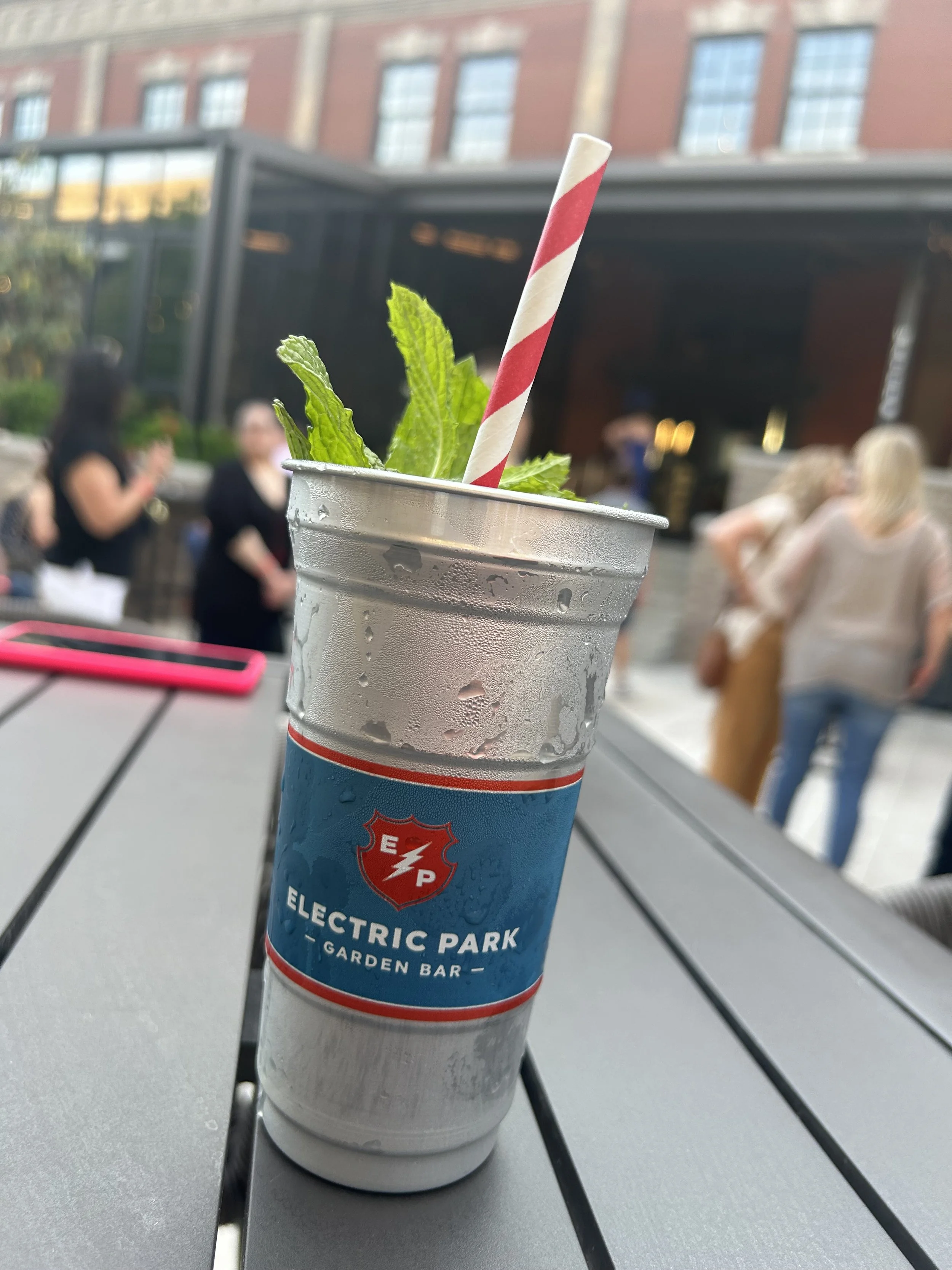 A cold beverage in an aluminum cup with a straw, and mint leaves, placed on a table outdoors at Electric Park Garden Bar with people in the background.