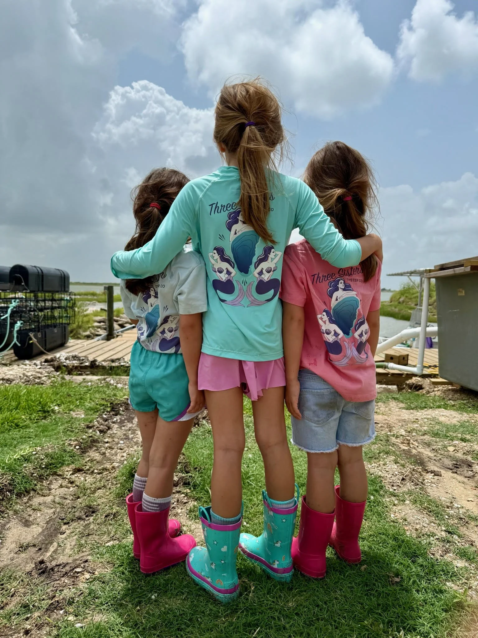 Three sisters wearing colorful rain boots and matching t-shirts with a mermaid design, standing close together with their arms around each other outdoors on a cloudy day.