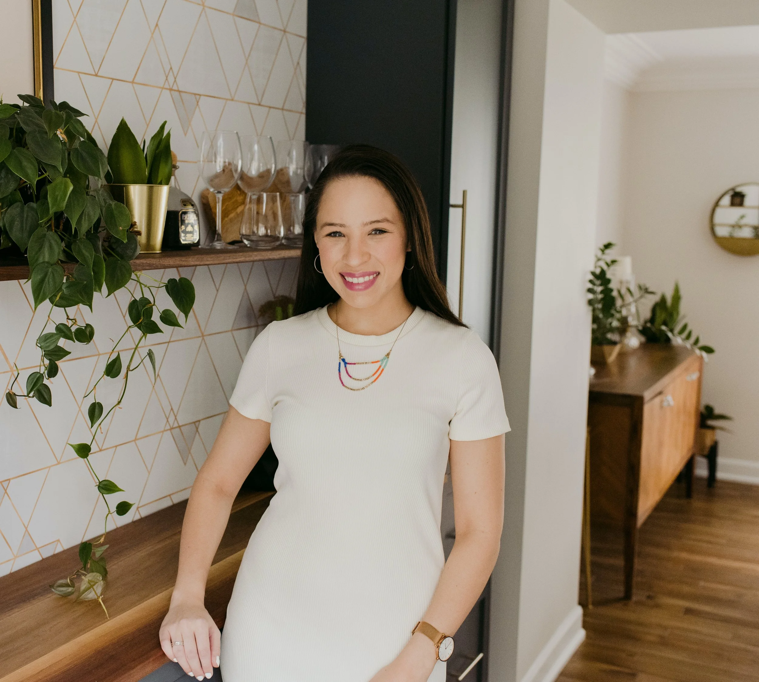 Smiling woman in white dress standing in modern living room with decorative wall, plants, and wooden furniture.