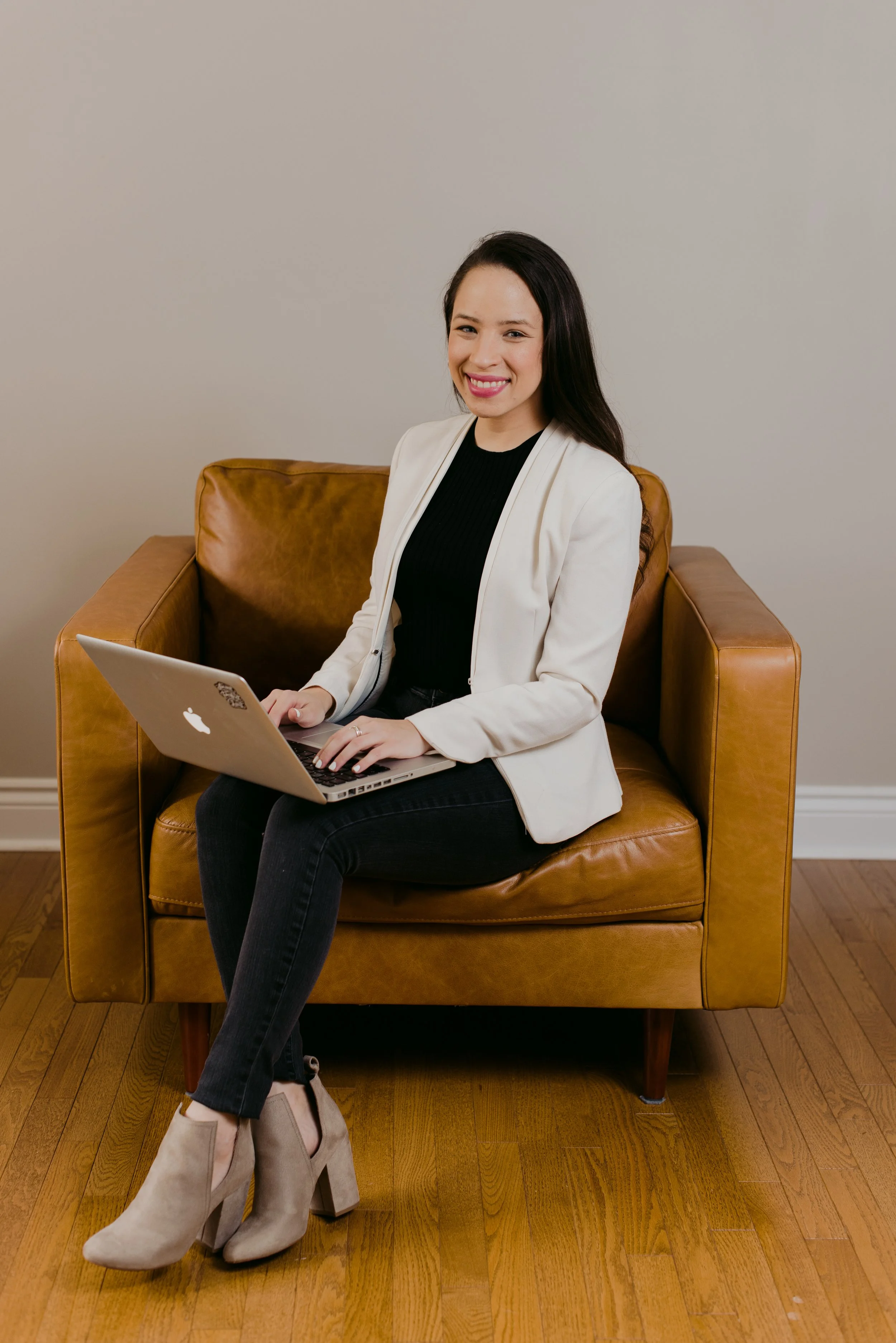 A woman with long dark hair smiling, sitting on a leather armchair, using a silver MacBook, in a room with hardwood floor and plain wall.