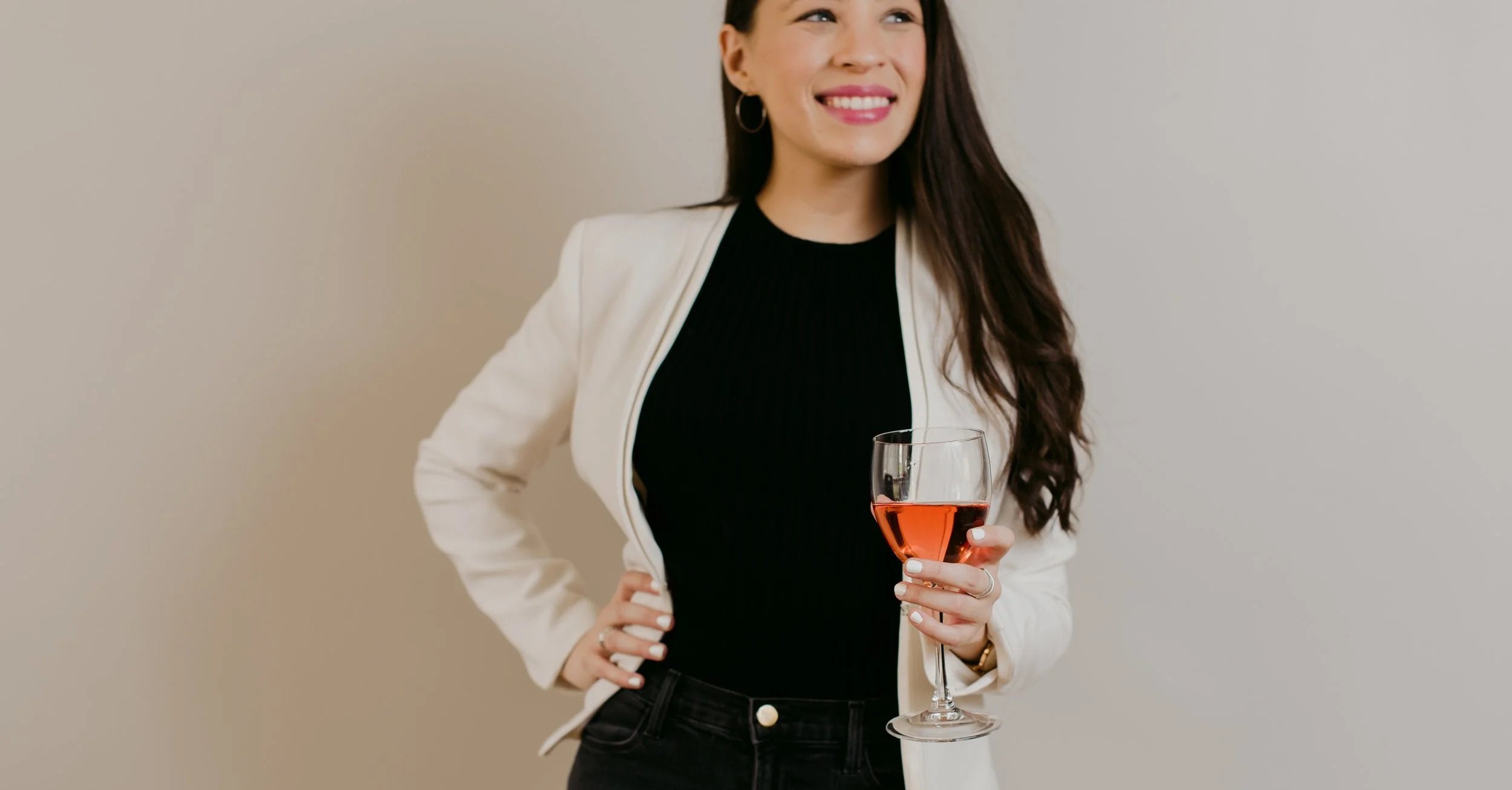 A woman in a white blazer and black top is holding a glass of rosé wine while smiling and standing against a plain light-colored wall.