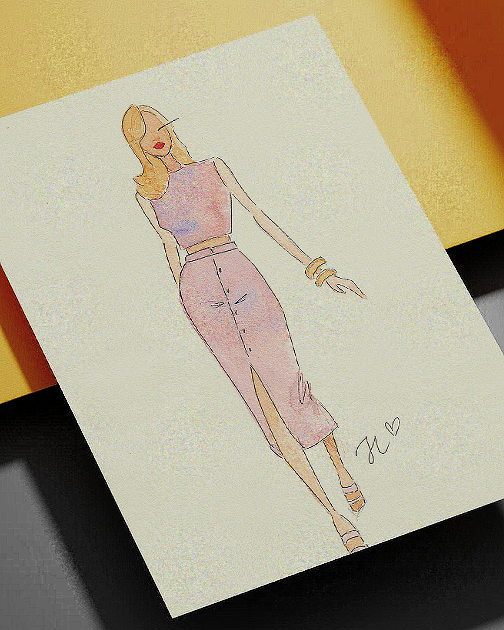 A fashion illustration of a woman in a sleeveless top and high-waisted, slit skirt, with blonde hair, gold bracelets, and high heels, signed with initials and a heart.