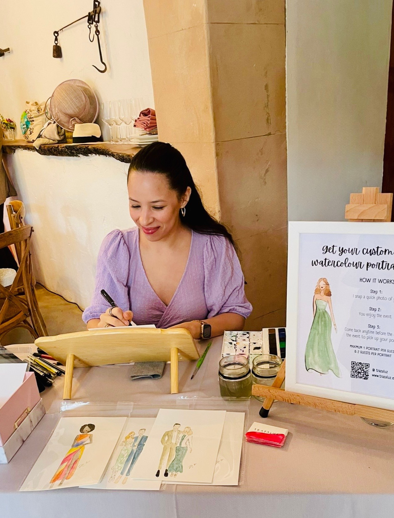 Woman in purple top sitting at table with watercolor portraits, signing artwork. Signage on easel promotes custom watercolor portraits with instructions.