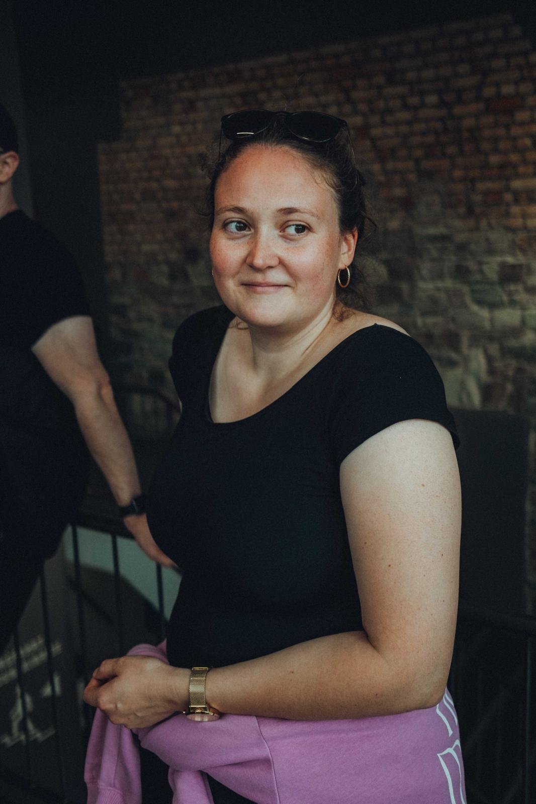 Junge Frau mit dunklem T-Shirt, rosa Pullover um die Taille und goldener Uhr, steht vor einer Steinmauer, schaut leicht zur Seite, trägt Sonnenbrille auf dem Kopf, goldene Ohrringe, lächelt leicht.