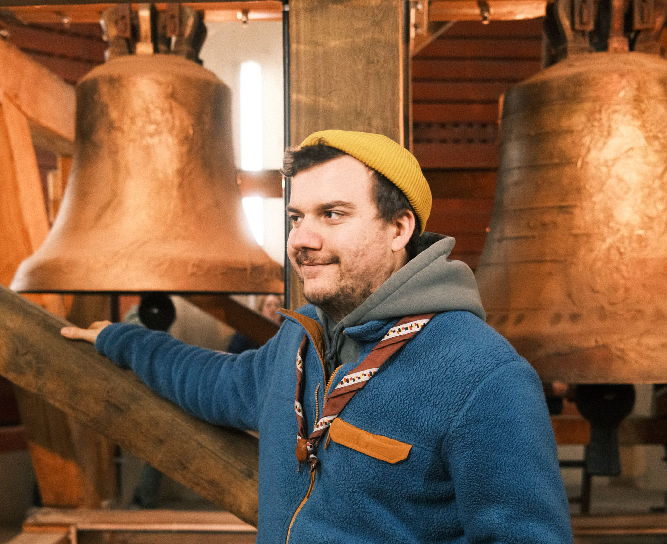 Ein junger Mann in einem blauen Jacke mit einer grauen Kapuze und gelbem Mütze, der eine große Glocke im Hintergrund berührt, in einem Holzhaus oder Museum.