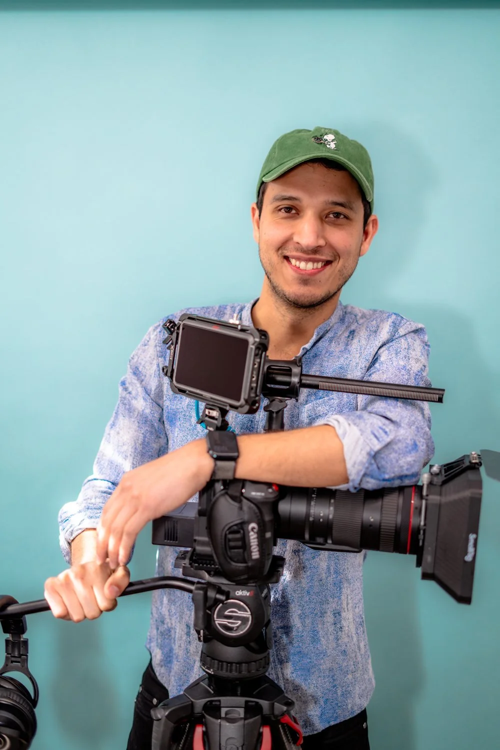 Hugo, wearing a blue shirt and green cap, smiling and leaning over a video camera
