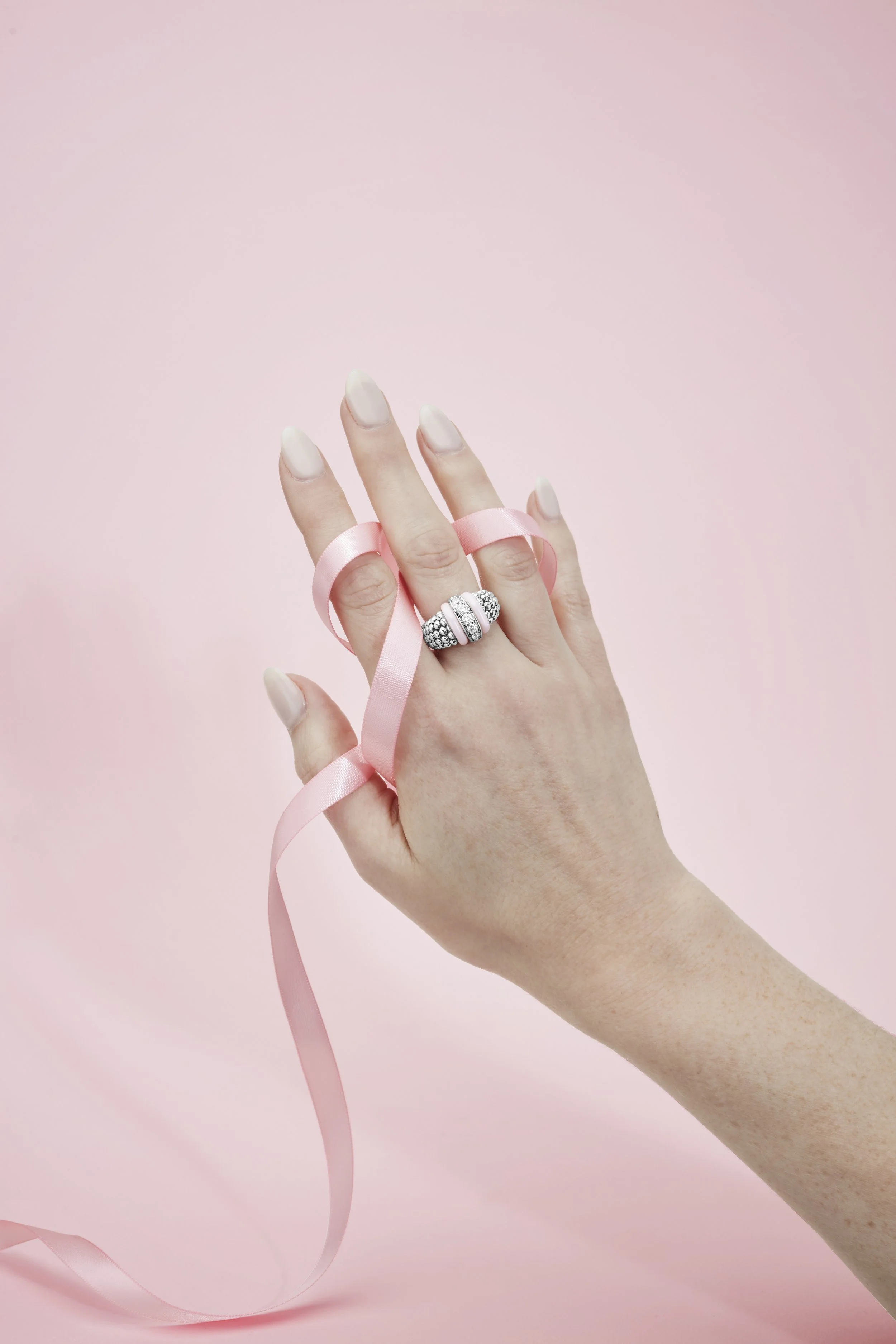 A person's left hand with a large, decorated ring on the ring finger, holding a pink ribbon against a soft pink background.