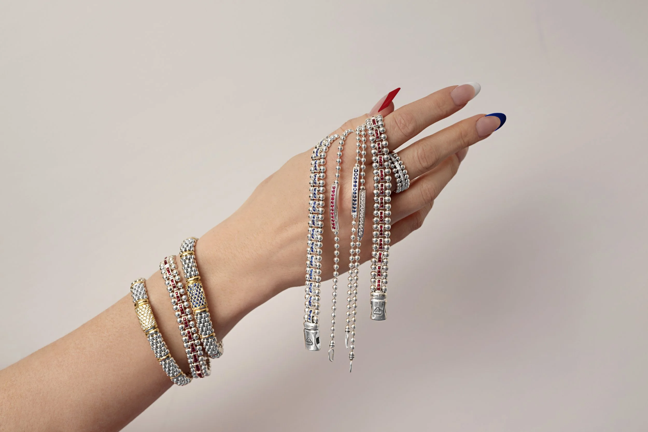 Hand displaying various silver and gold jewelry including bracelets, rings, and necklaces with red, blue, and clear gemstones against a plain background.
