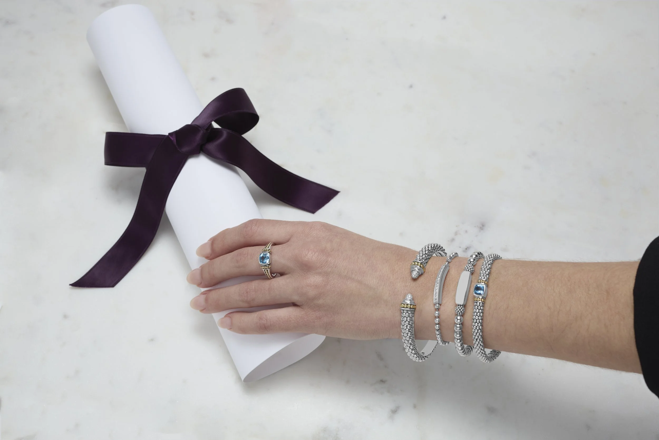 A hand with silver and gold jewelry resting on a white surface with a rolled-up white paper tied with a dark purple ribbon.