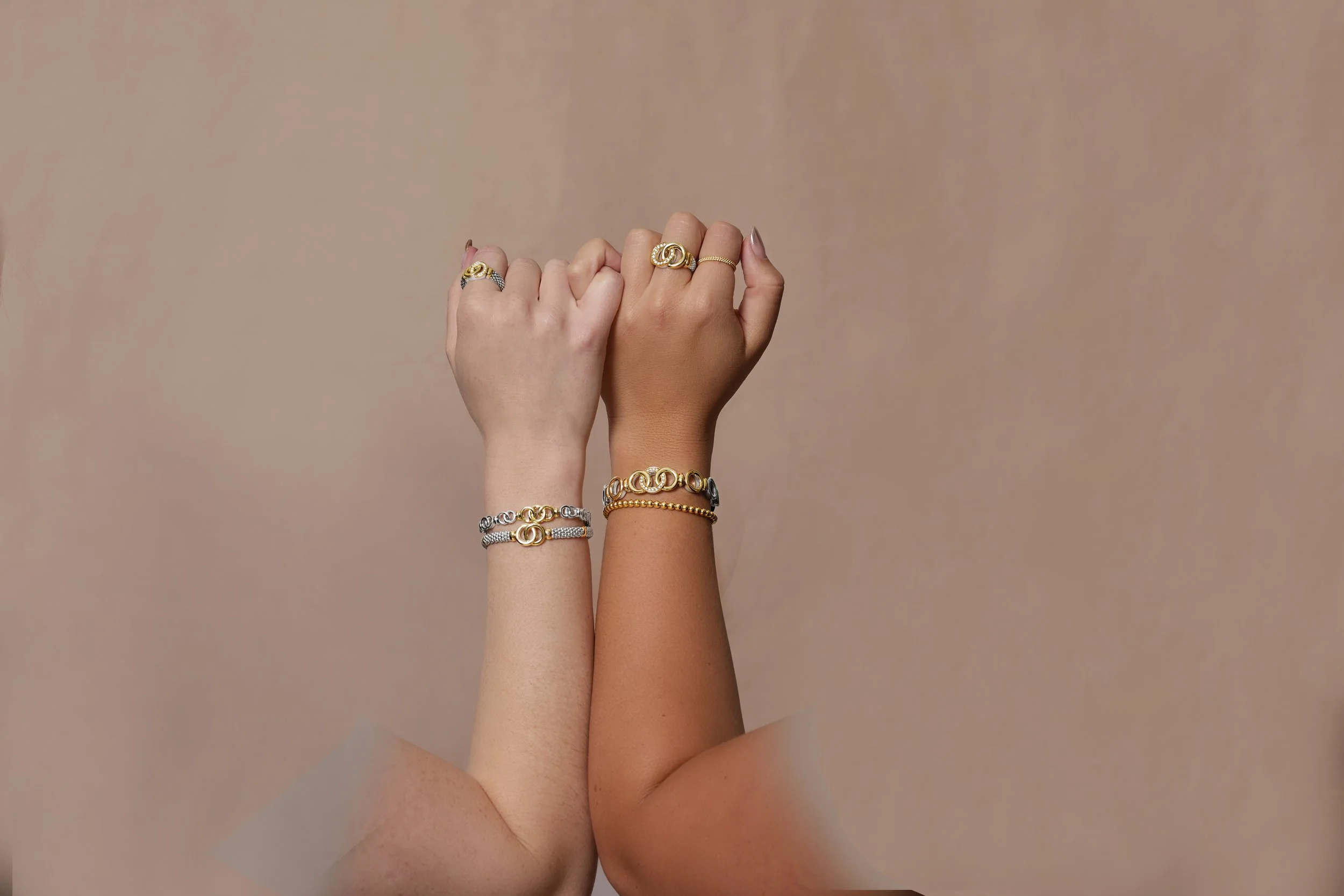 Two hands with bracelets and rings, showing a gesture of strength or solidarity, against a plain beige background.