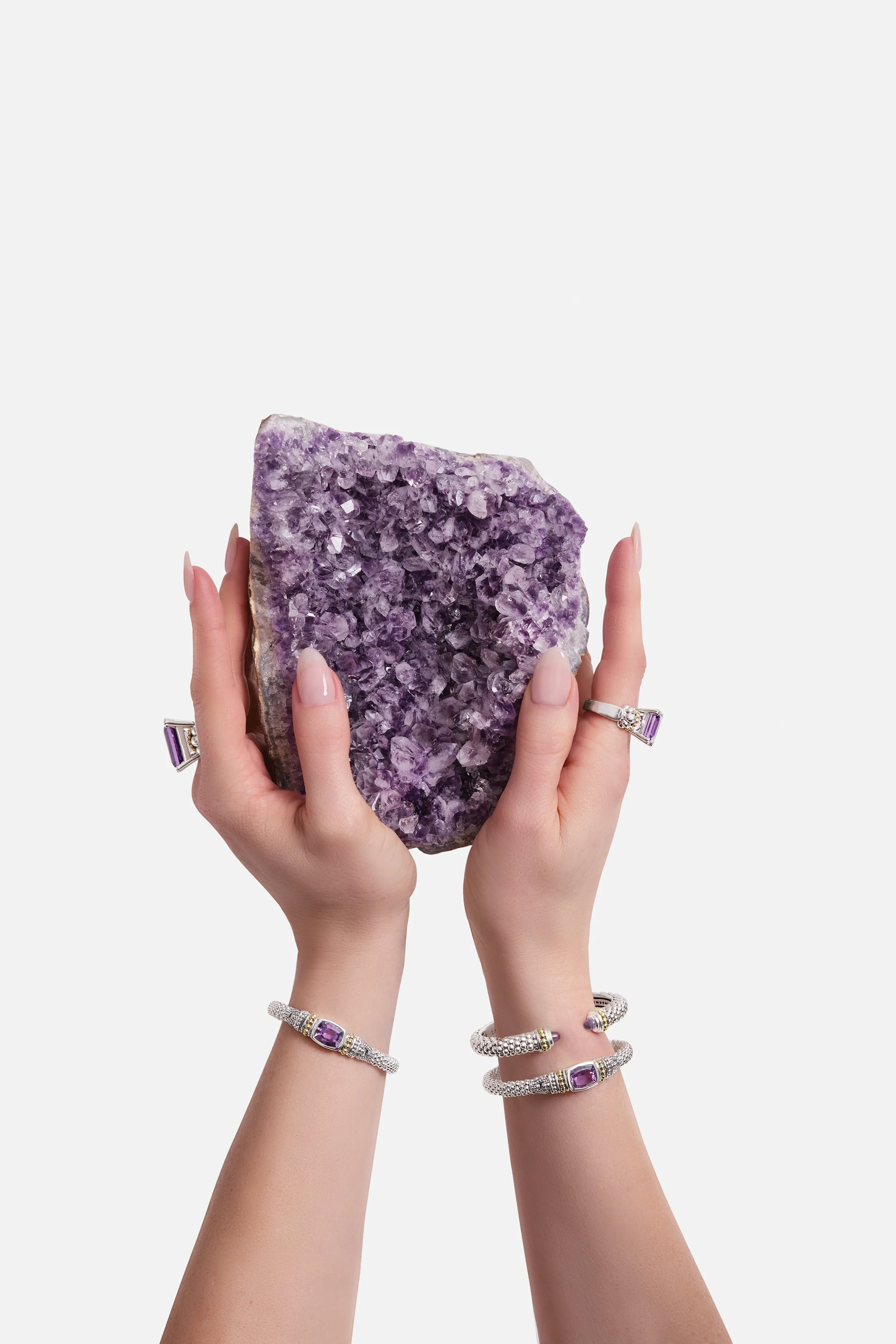 Hands holding a large purple geode gemstone, adorned with rings and bracelets featuring purple gemstones.