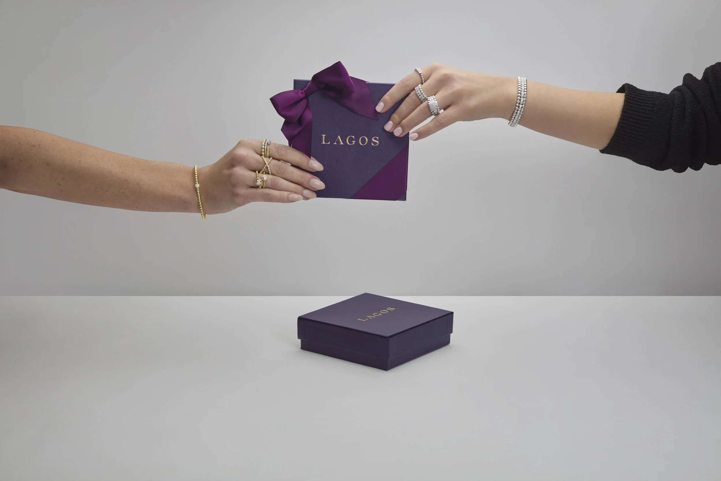 Two hands exchanging a purple gift box with gold lettering 'LAGOS', one hand with gold jewelry and rings, and the other with silver jewelry and rings, over a white surface with a matching purple box below.