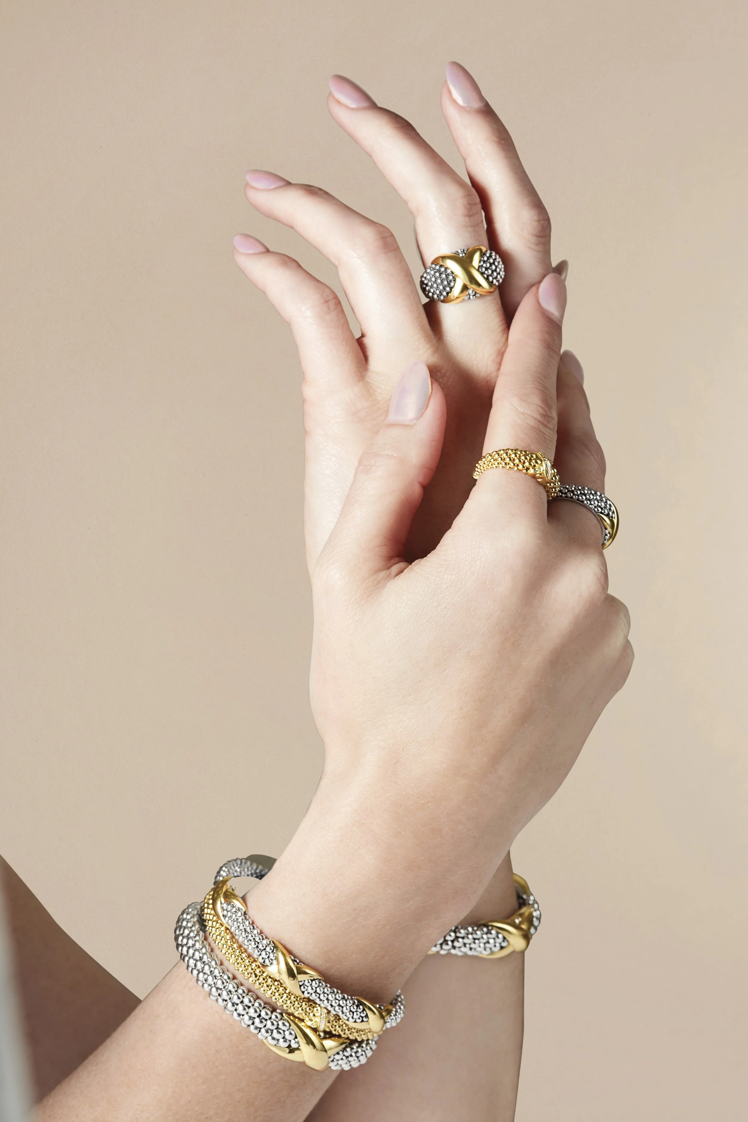 A person's hands and wrists adorned with multiple gold and silver rings and bangles, posed against a beige background.