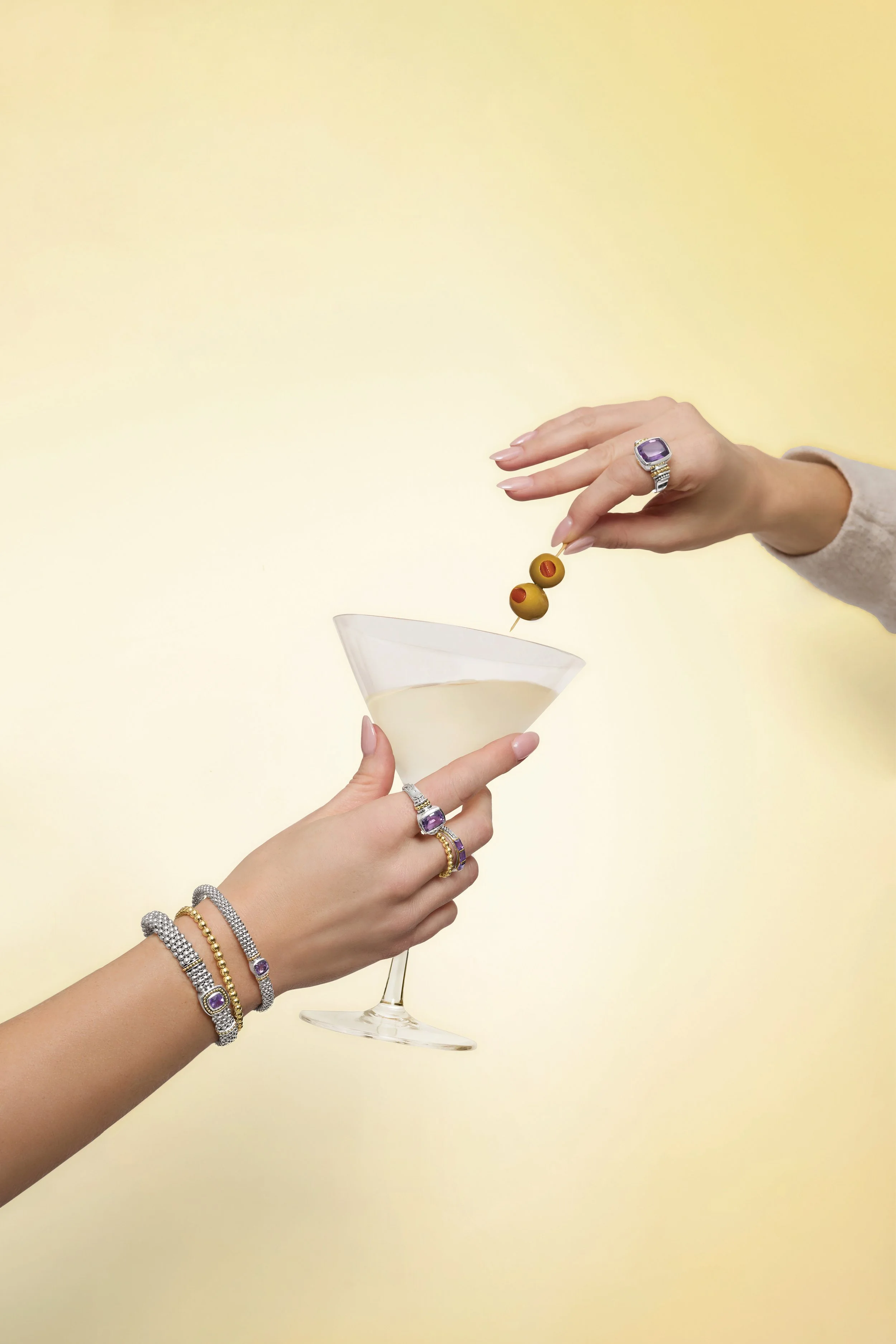 Two women exchanging a drink, one holding a martini glass with olives and the other about to add more olives, all wearing rings and bracelets.