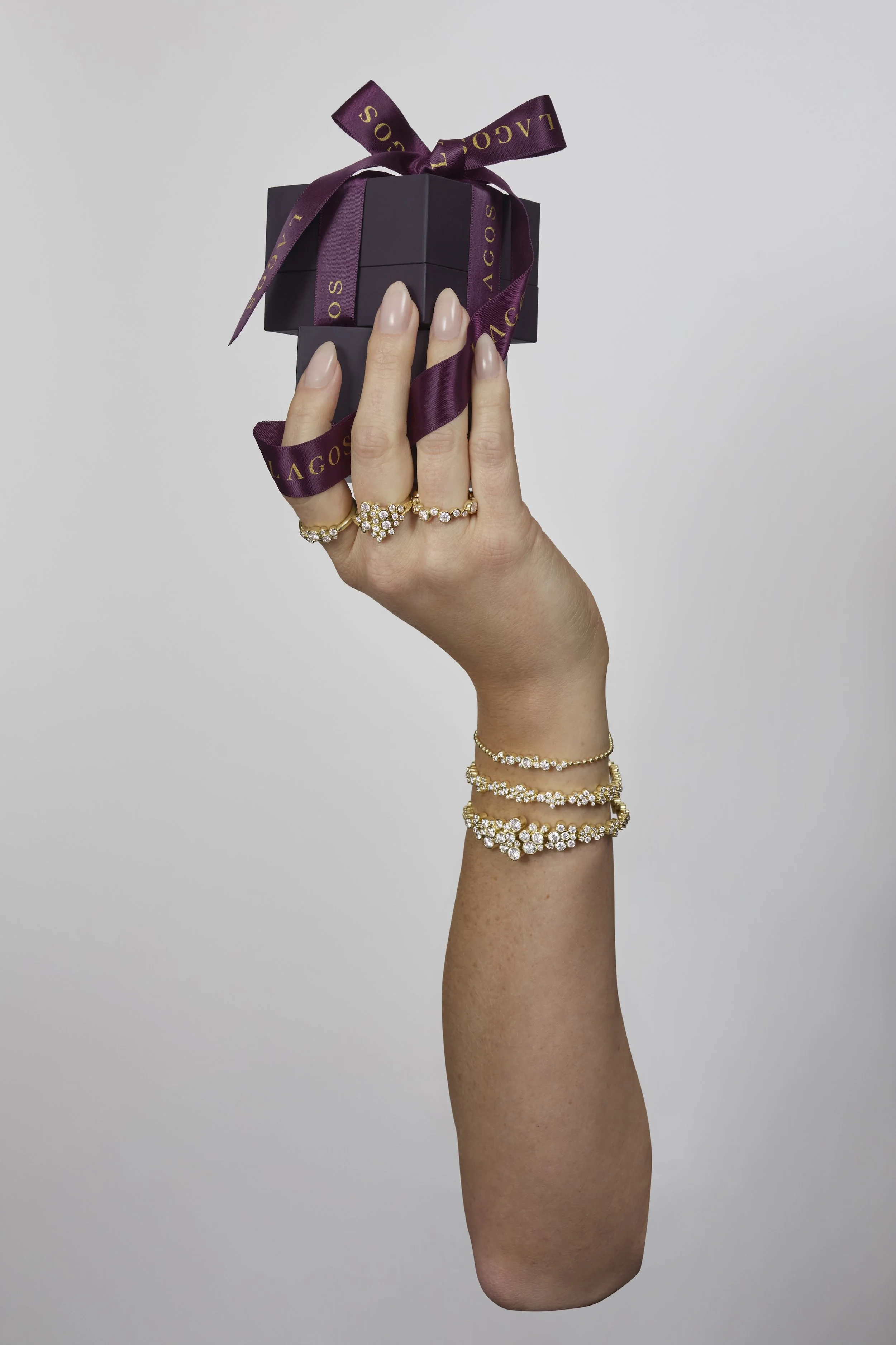 Hand with multiple jewelry rings and bracelets holding a small purple gift box with a purple ribbon, against a plain gray background.