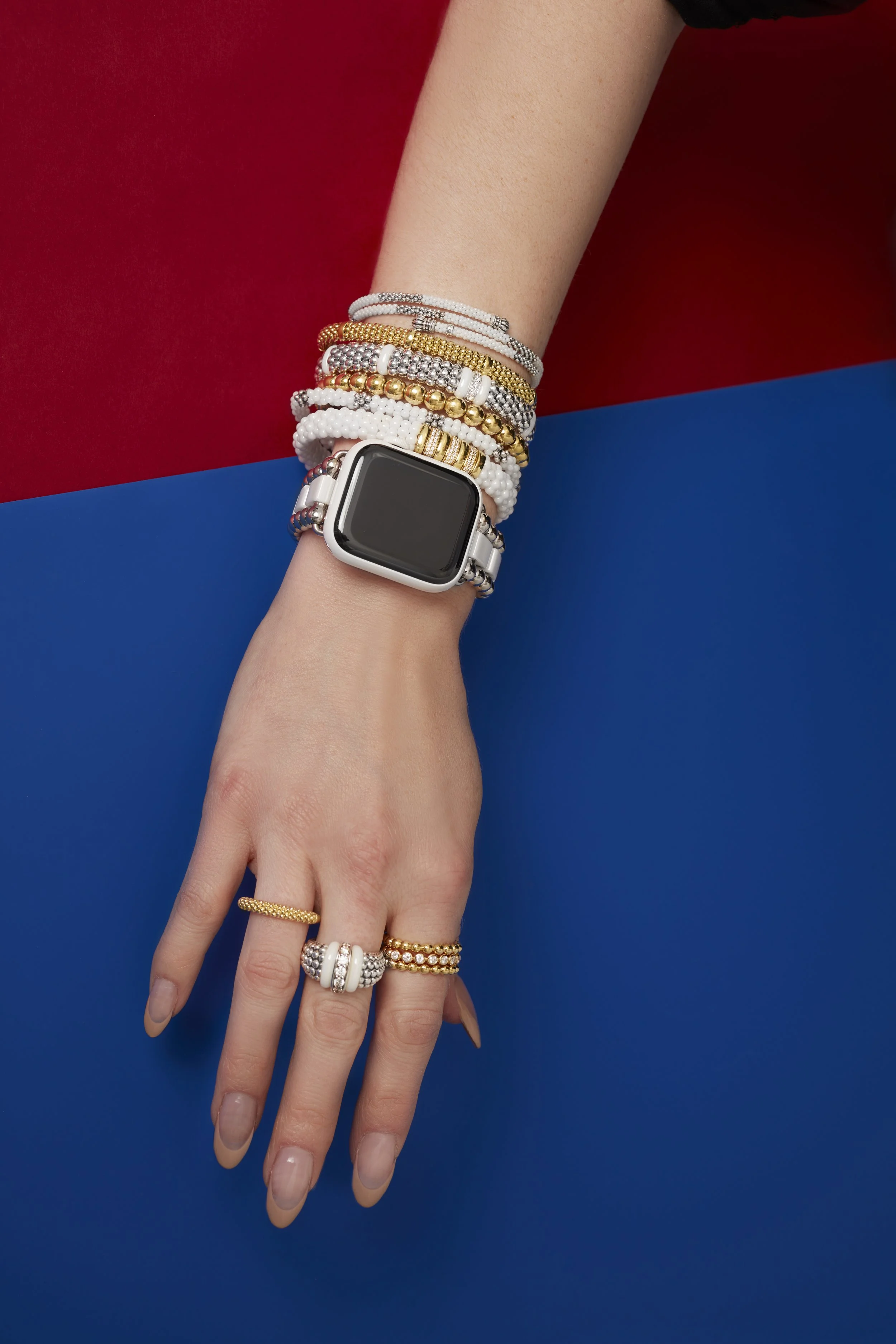 A person's arm and hand with multiple gold and white beaded bracelets and rings, wearing a smartwatch on the wrist, against a background divided into red and blue sections.