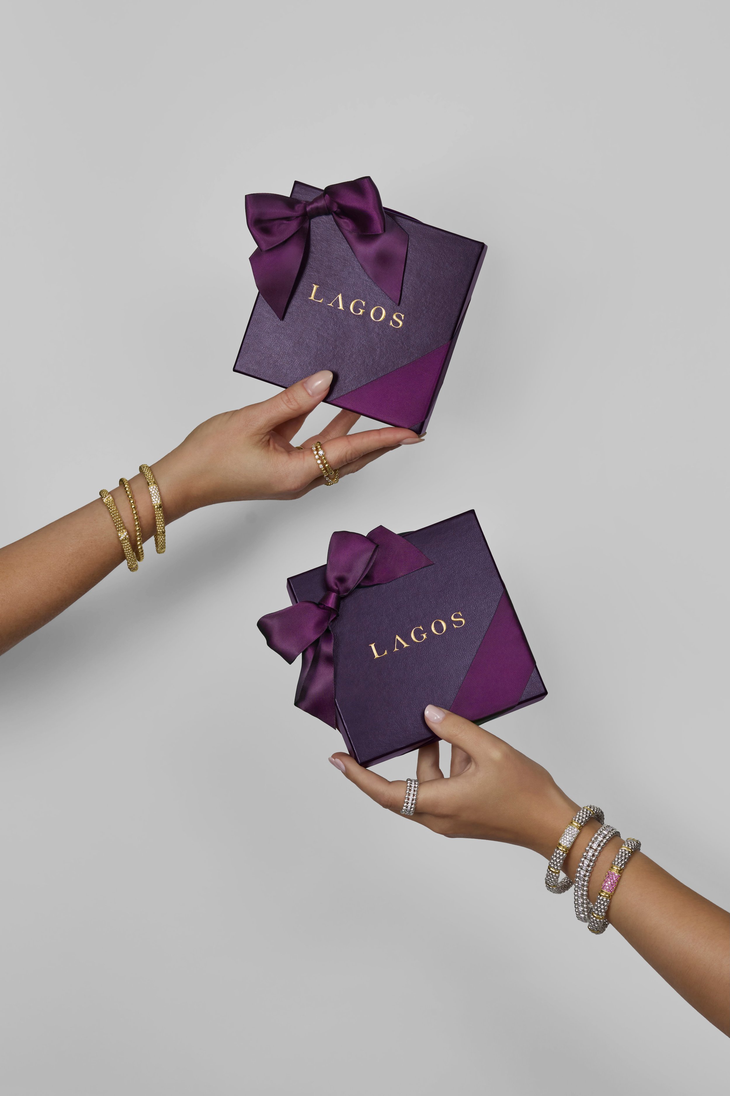 Two hands holding purple gift boxes with gold 'LAGOS' branding and purple satin bows on a gray background.