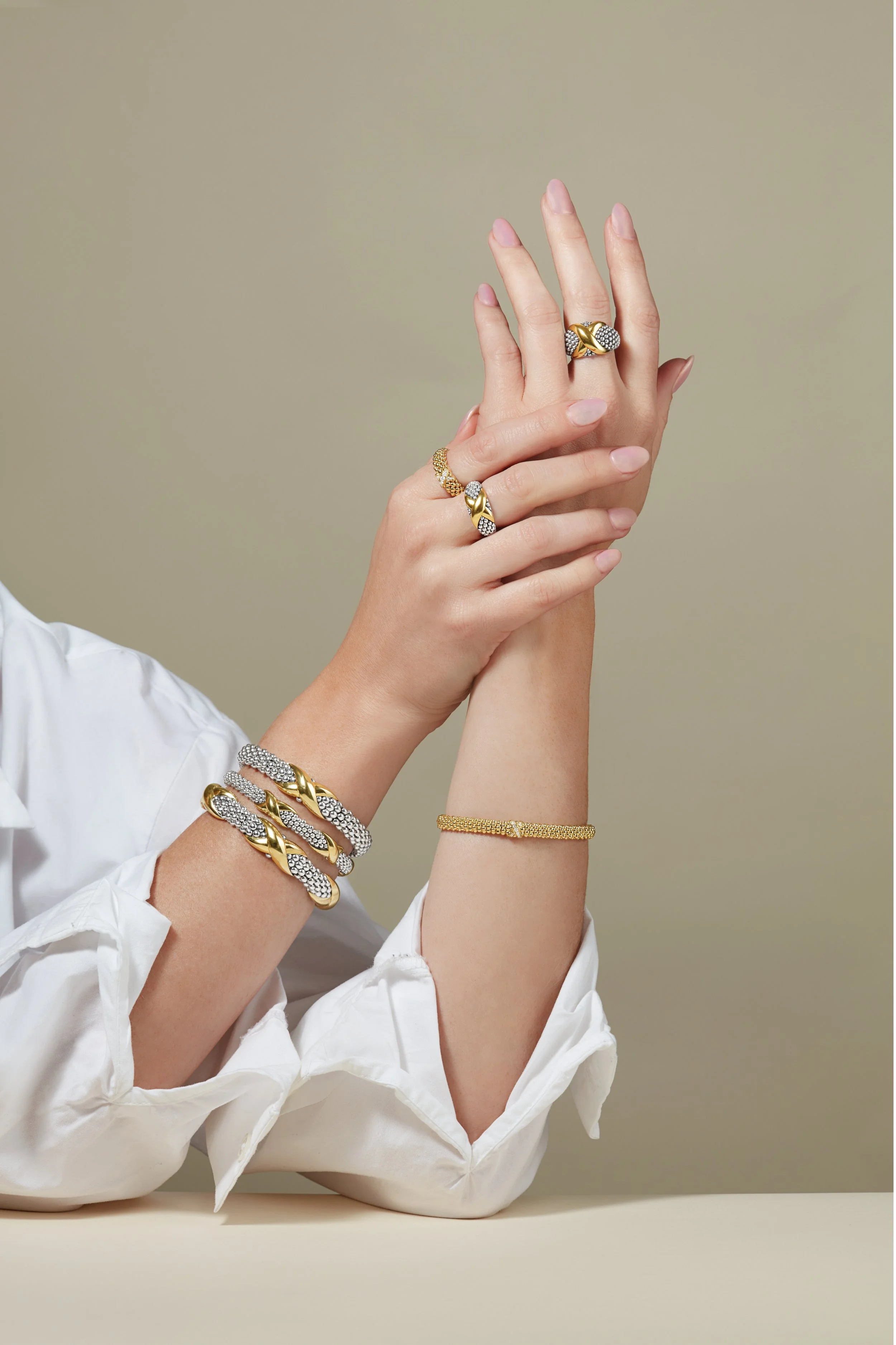 Close-up of a woman's hands and wrists adorned with gold and diamond jewelry, including rings, bracelets, and a white shirt sleeve, against a neutral background.